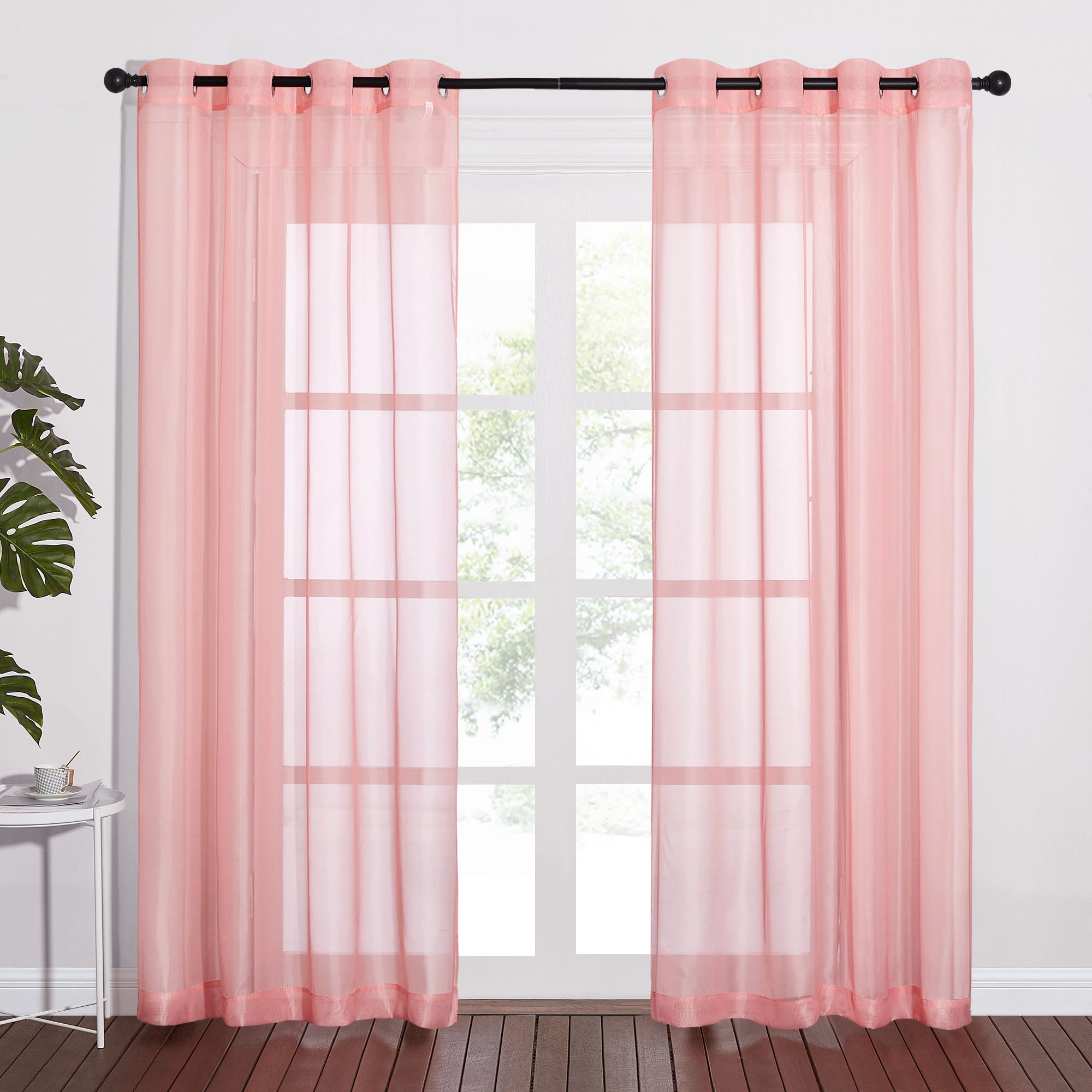 Two light pink sheer curtains with metal grommets hanging on a black rod, filtering light from a window.