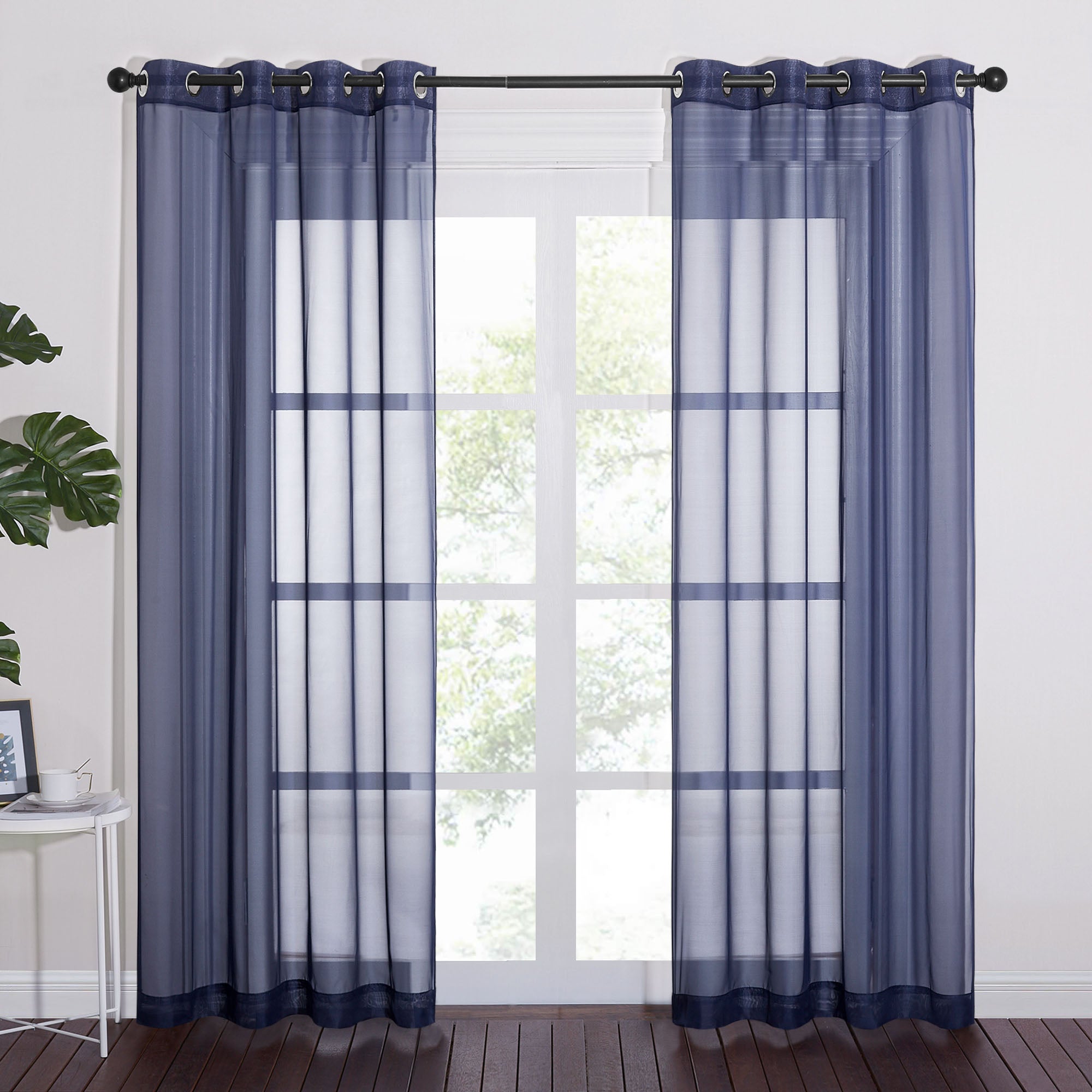 Two navy blue light sheer curtains with grommet tops hanging on a rod, filtering natural light from a tall window.