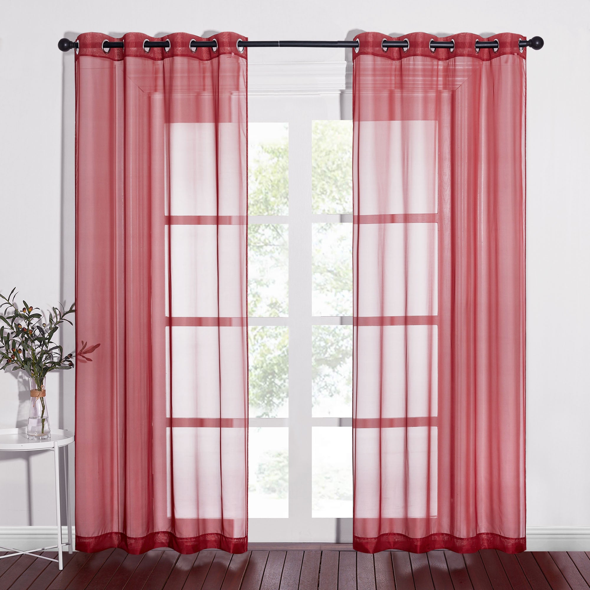 Two red sheer curtains with silver grommets hang on a black curtain rod, filtering light into a room with wood floors.