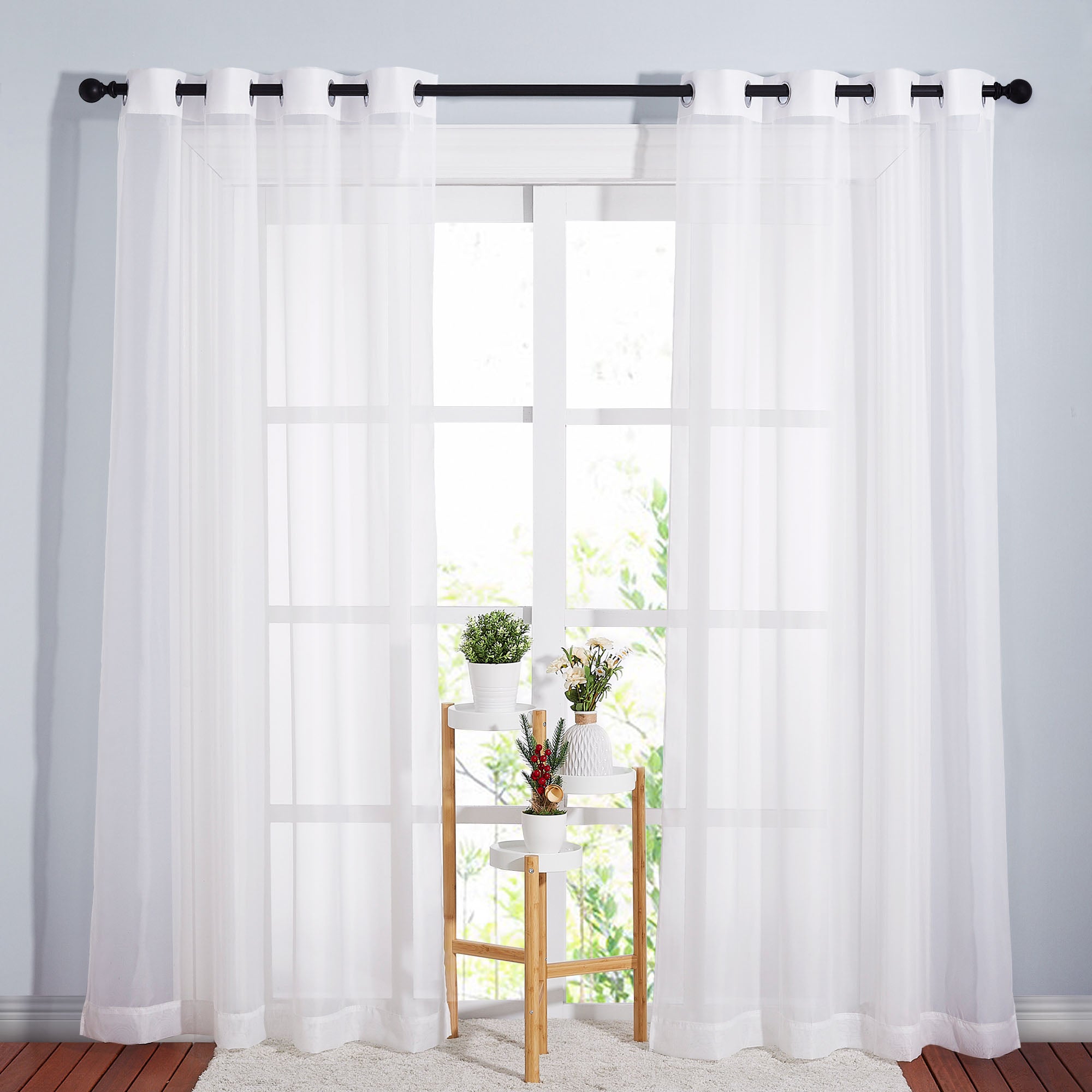 Full-length white sheer curtains with grommets hang on a rod, filtering light from a large window with a plant stand.