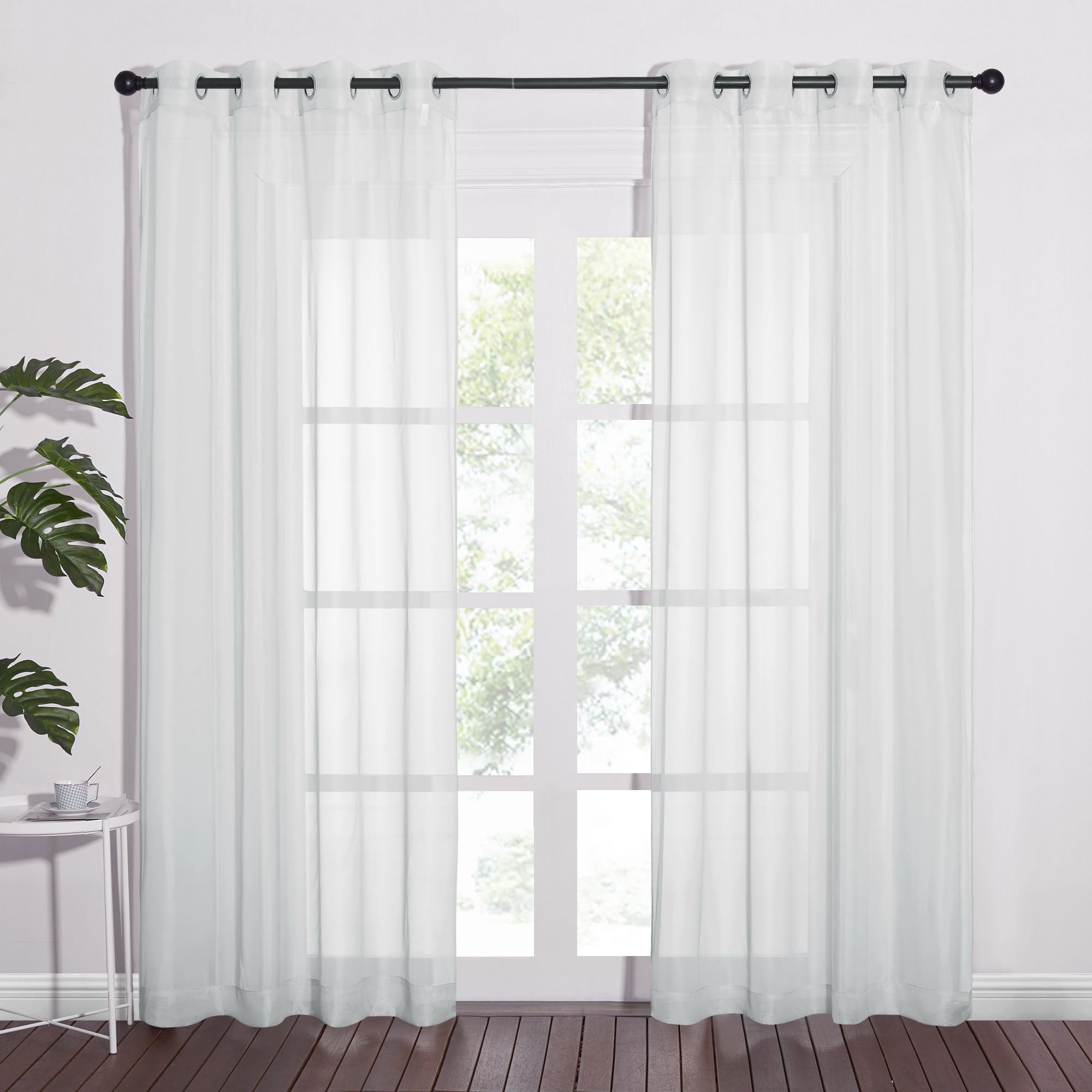 White sheer grommet-top curtains hanging on a dark rod over a large window in a room with a wood floor.