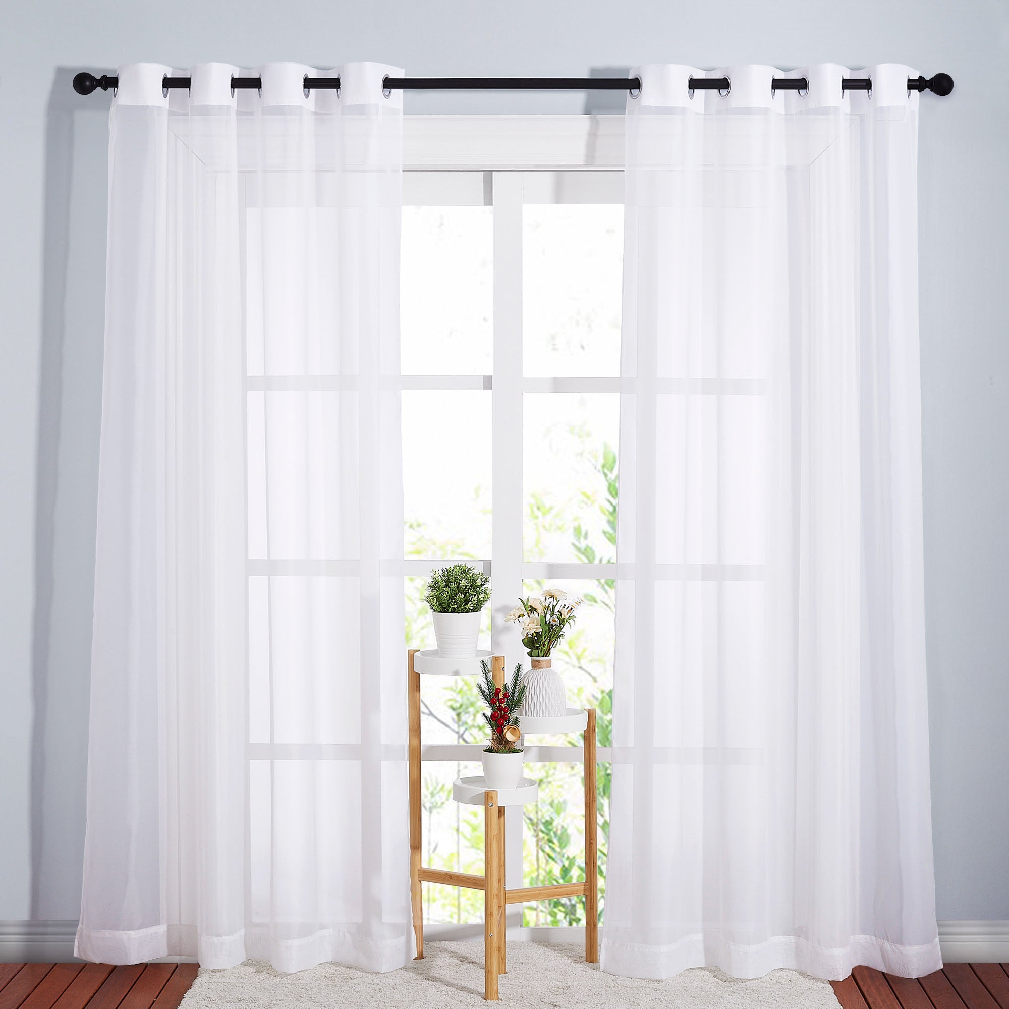 White sheer curtains with silver grommets hanging on a black rod, letting light in through a window with a plant stand.
