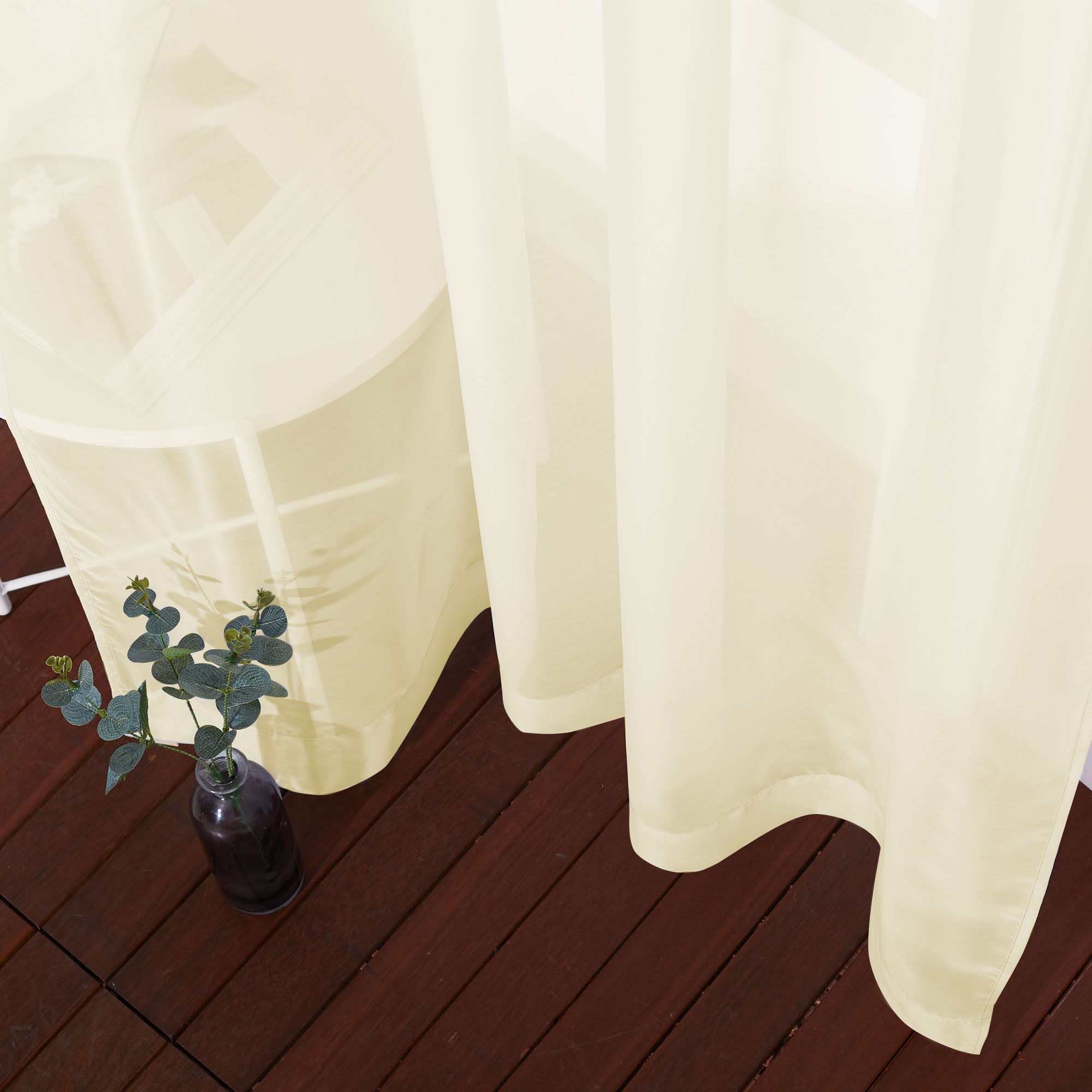 High-angle view of a light sheer curtain's translucent fabric and bottom hem draping over a dark wood floor.