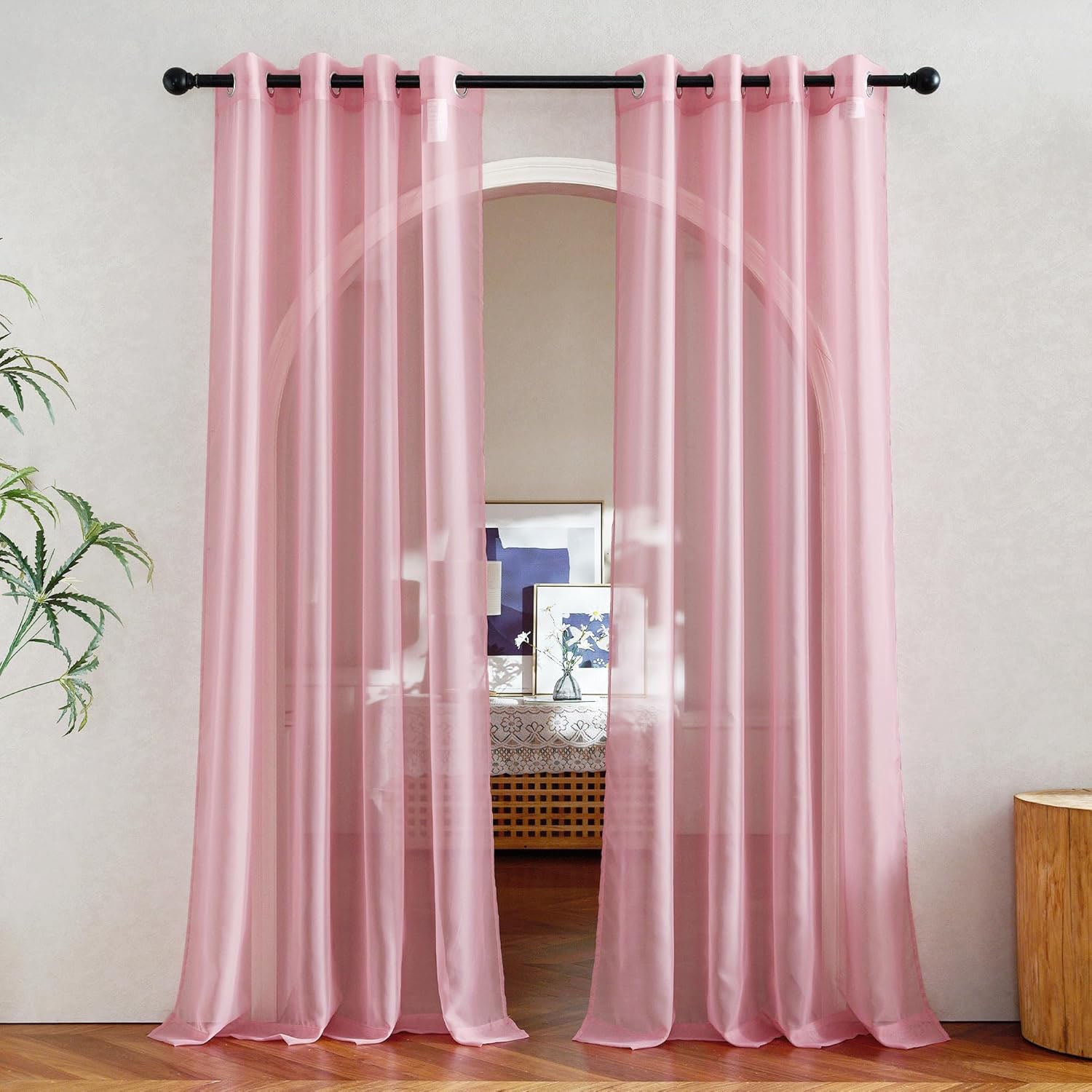 A pair of light pink sheer curtains with silver grommets hanging on a black rod, framing an arched window in a modern room.