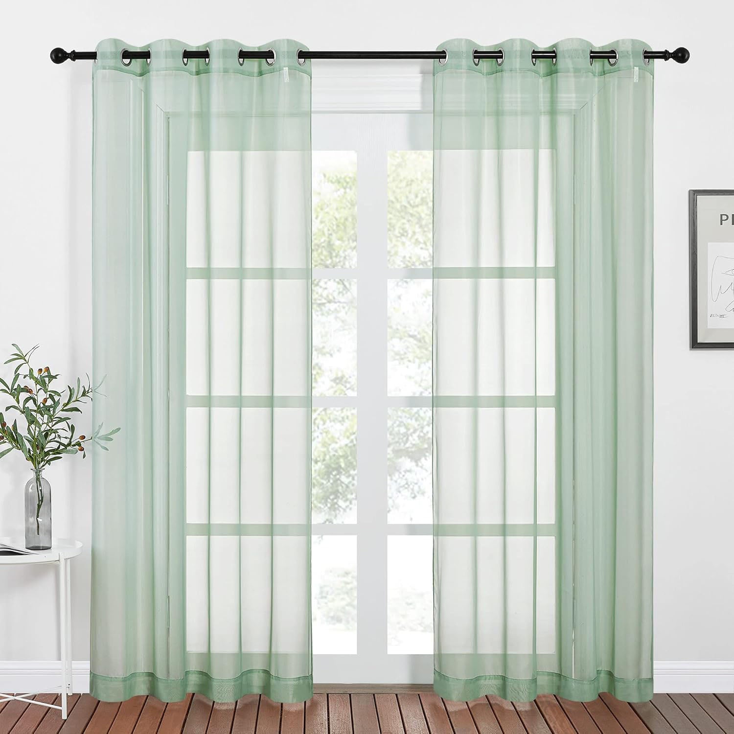 Pair of light sage green sheer curtains with silver grommet tops hanging on a black rod in front of a tall window.
