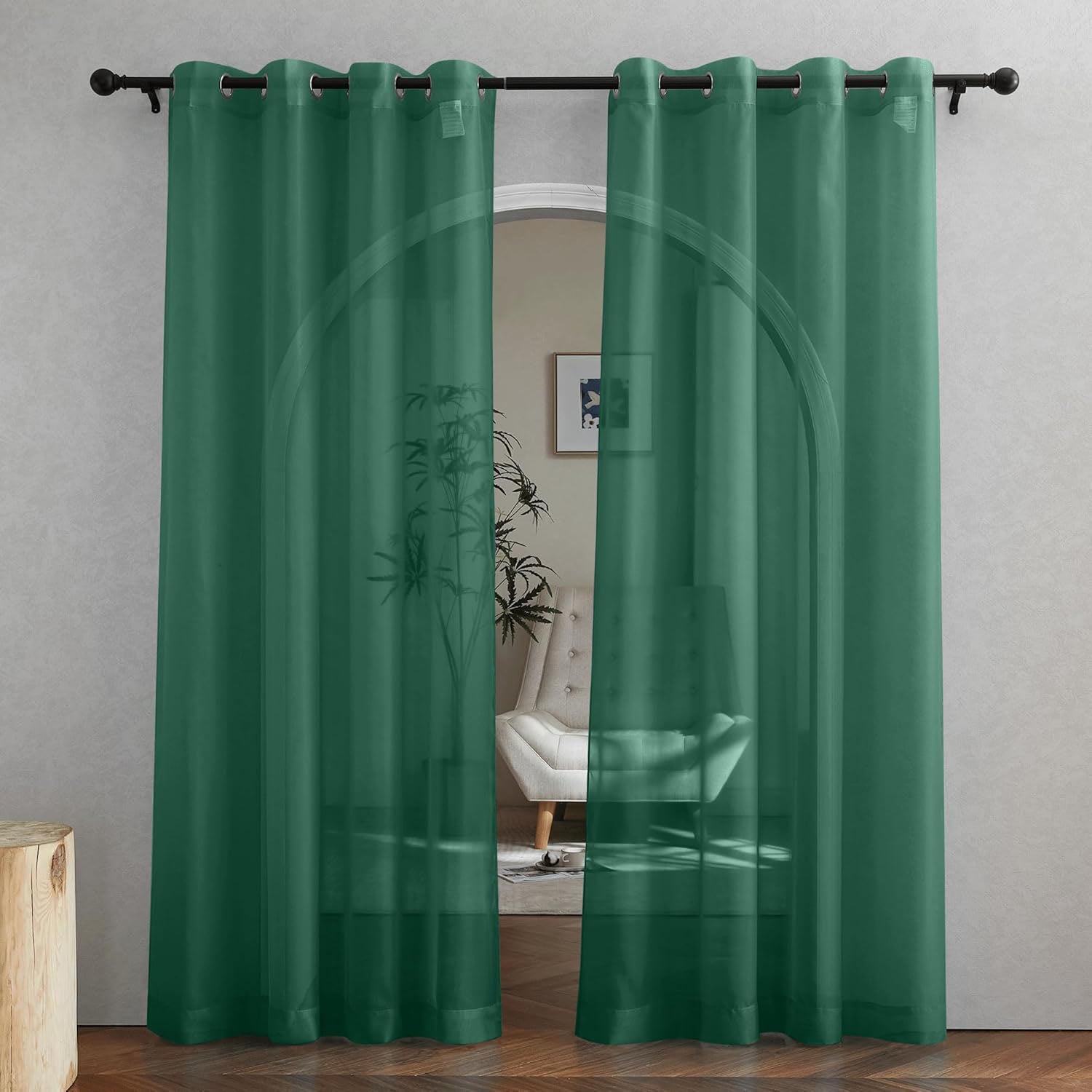 Two dark green sheer curtain panels with silver grommets hang on a black rod, revealing a room through the fabric.