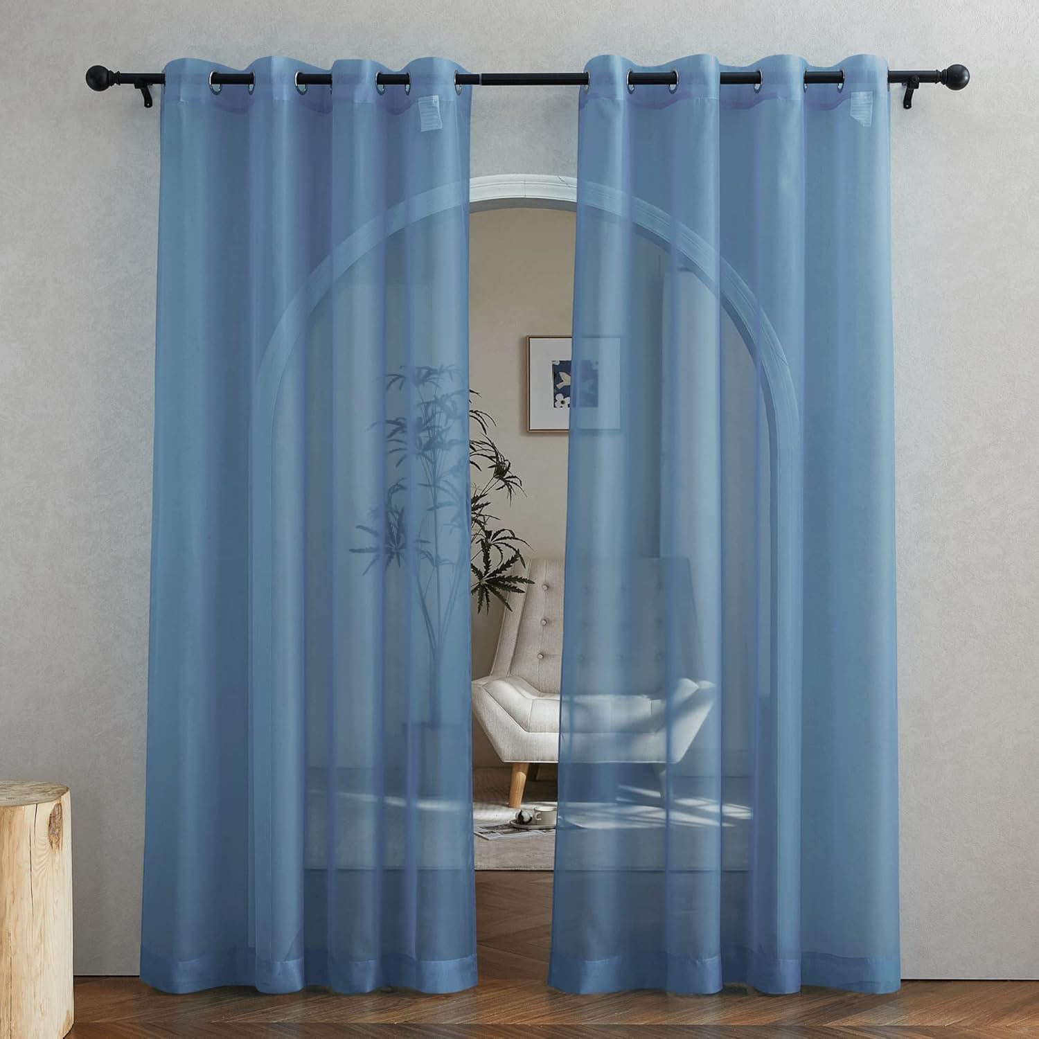 A pair of dusty blue sheer curtains with grommets hanging on a black rod, filtering light in front of an arched doorway.