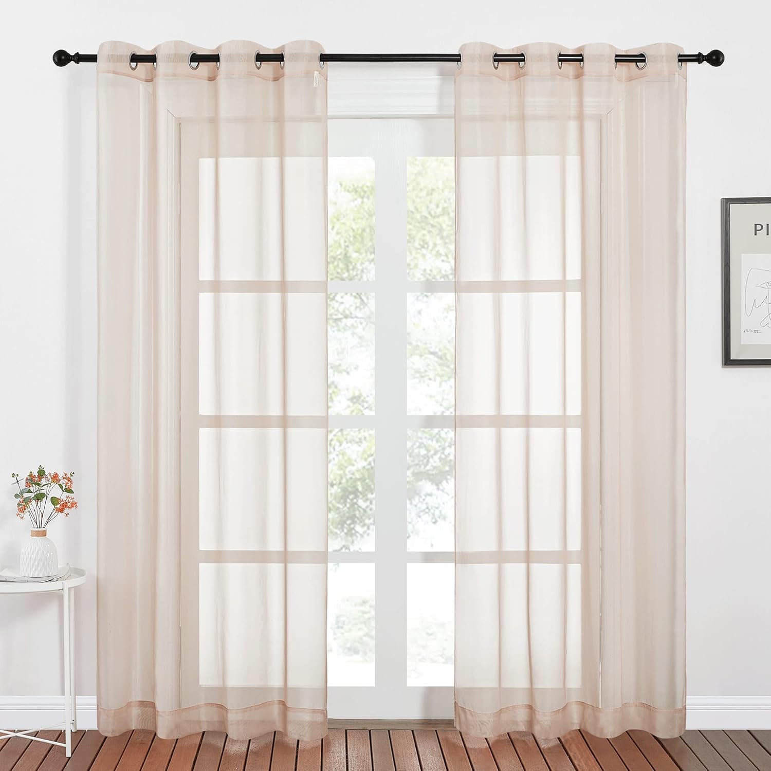 Two light beige sheer curtain panels with silver grommets hang on a black rod, filtering light from a large window.