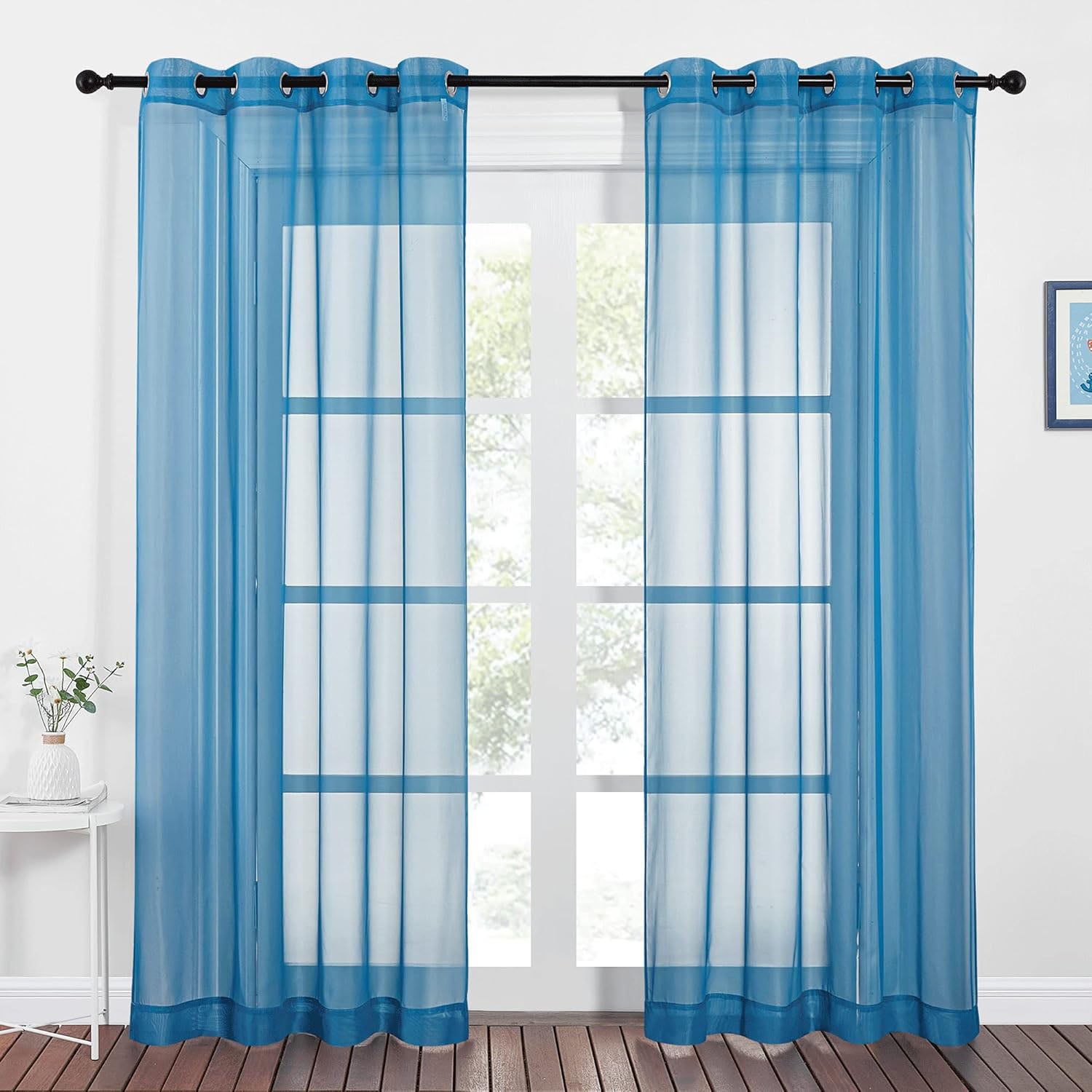 A pair of sheer blue grommet-top curtains hanging on a black rod, filtering daylight from a large window.