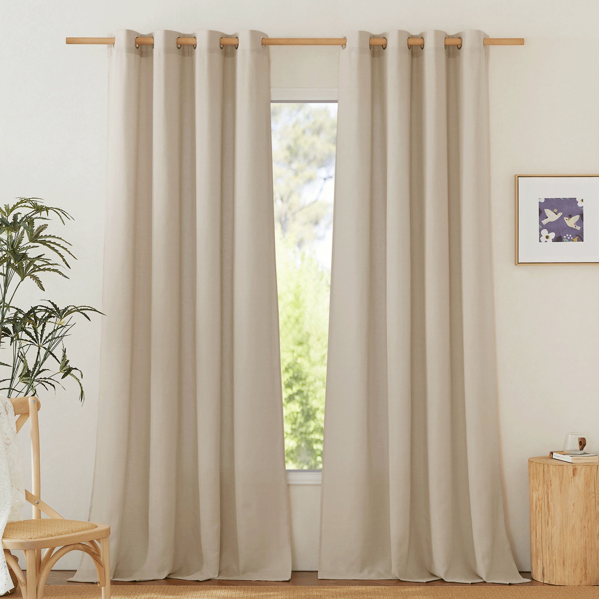 Full view of beige linen curtains with grommets hanging on a wooden rod over a window in a brightly lit, neutral room.