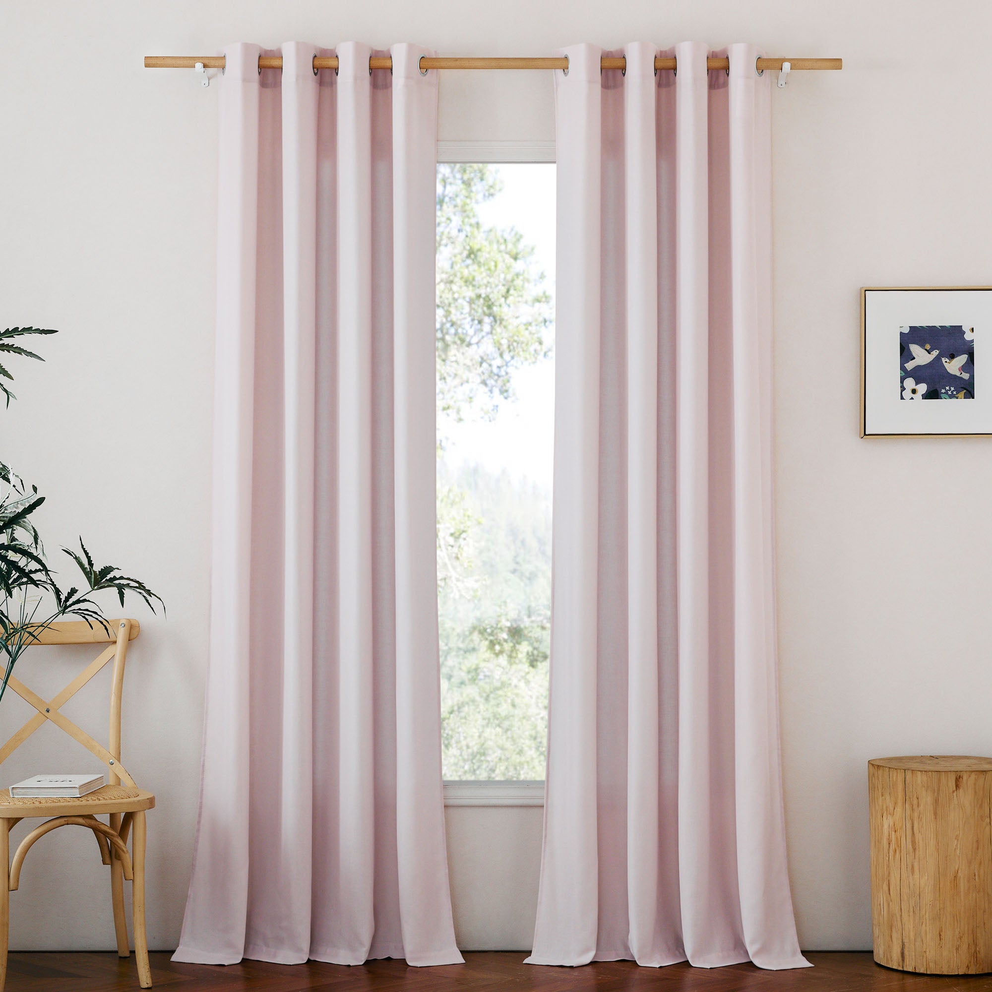 Light pink linen curtains with silver grommets hang on a wooden rod, framing a window in a naturally styled room.