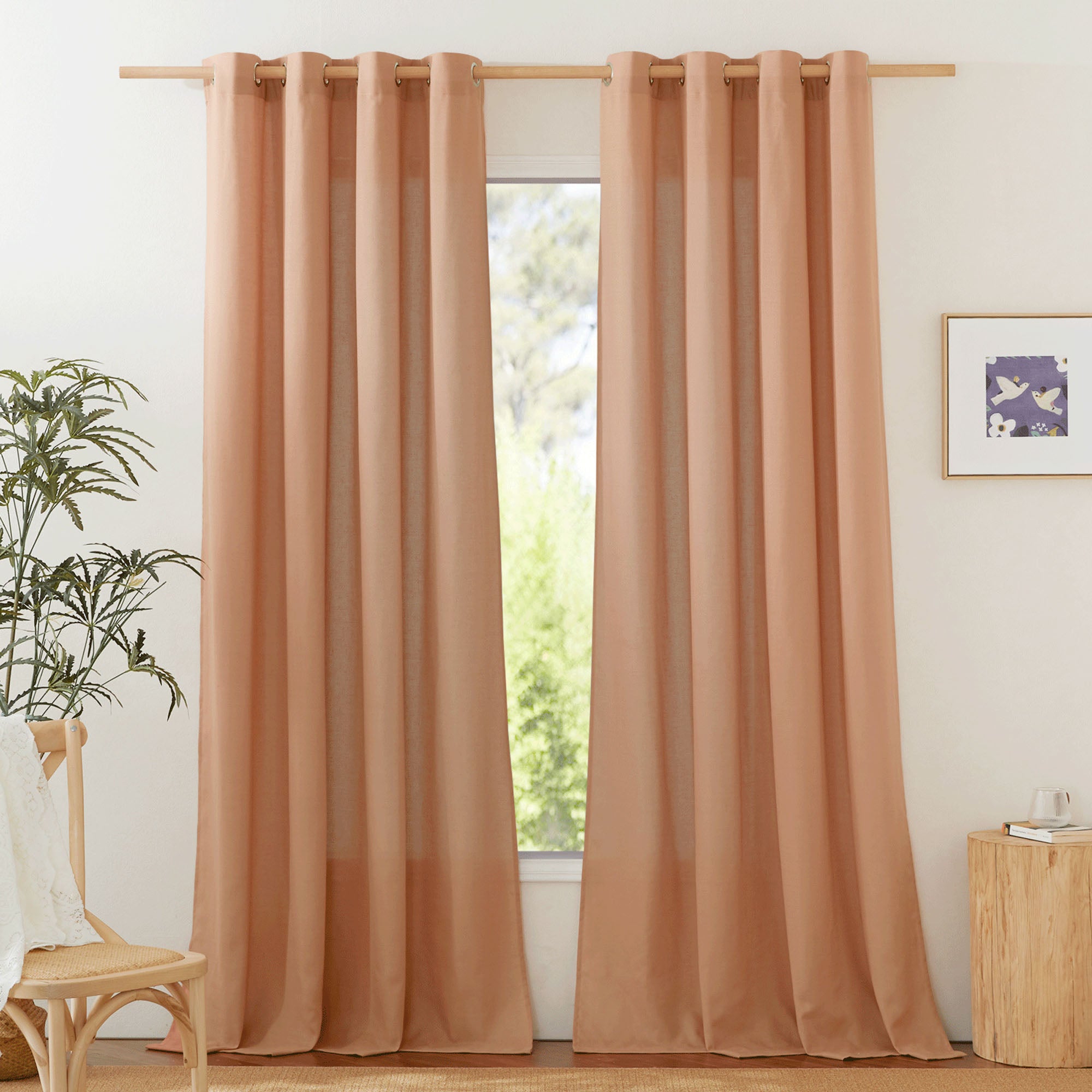 A pair of terracotta linen curtains with silver grommets hanging on a wooden rod, framing a window in a decorated room.