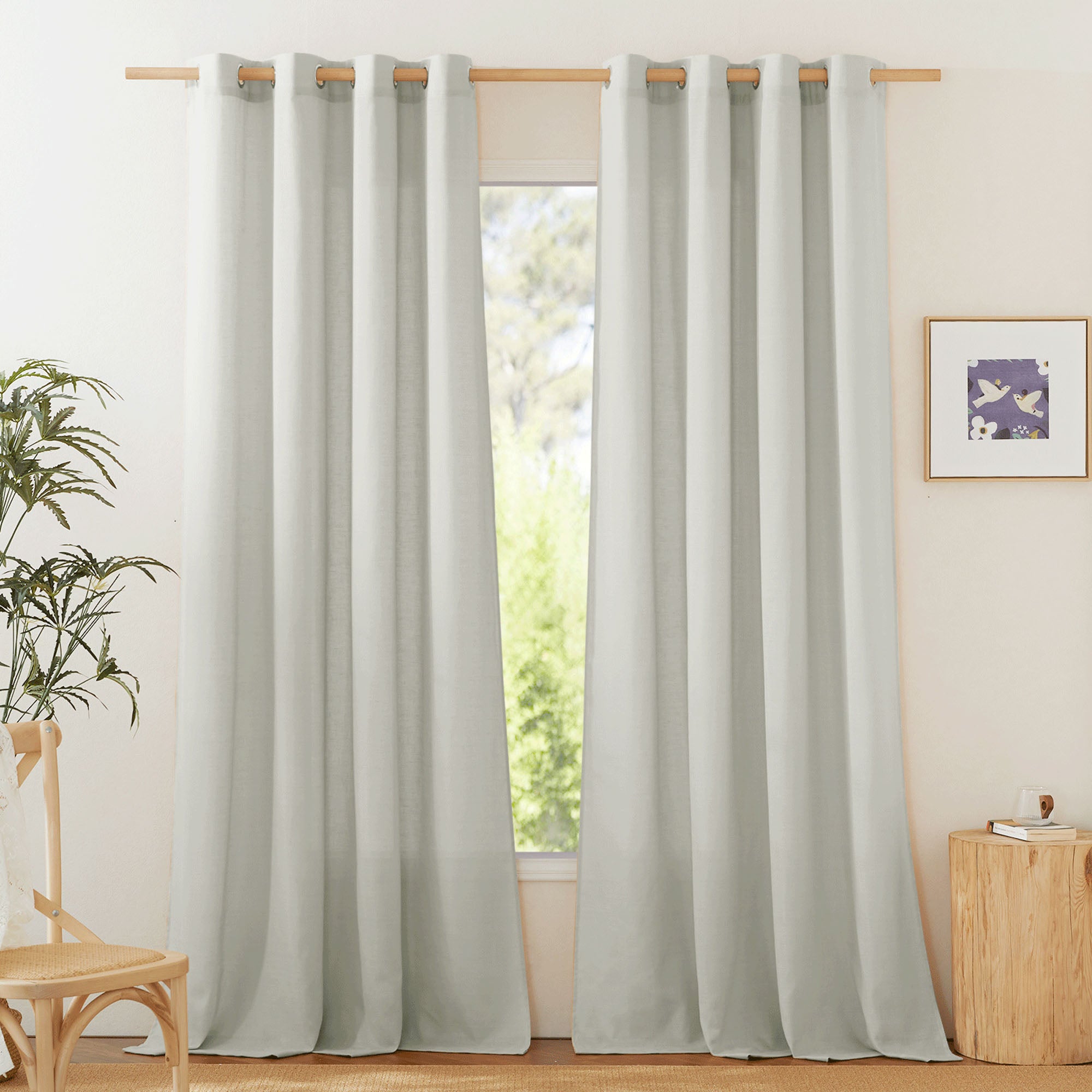Two light gray linen curtain panels with silver grommets hanging on a wooden rod, shown in a sunlit room.