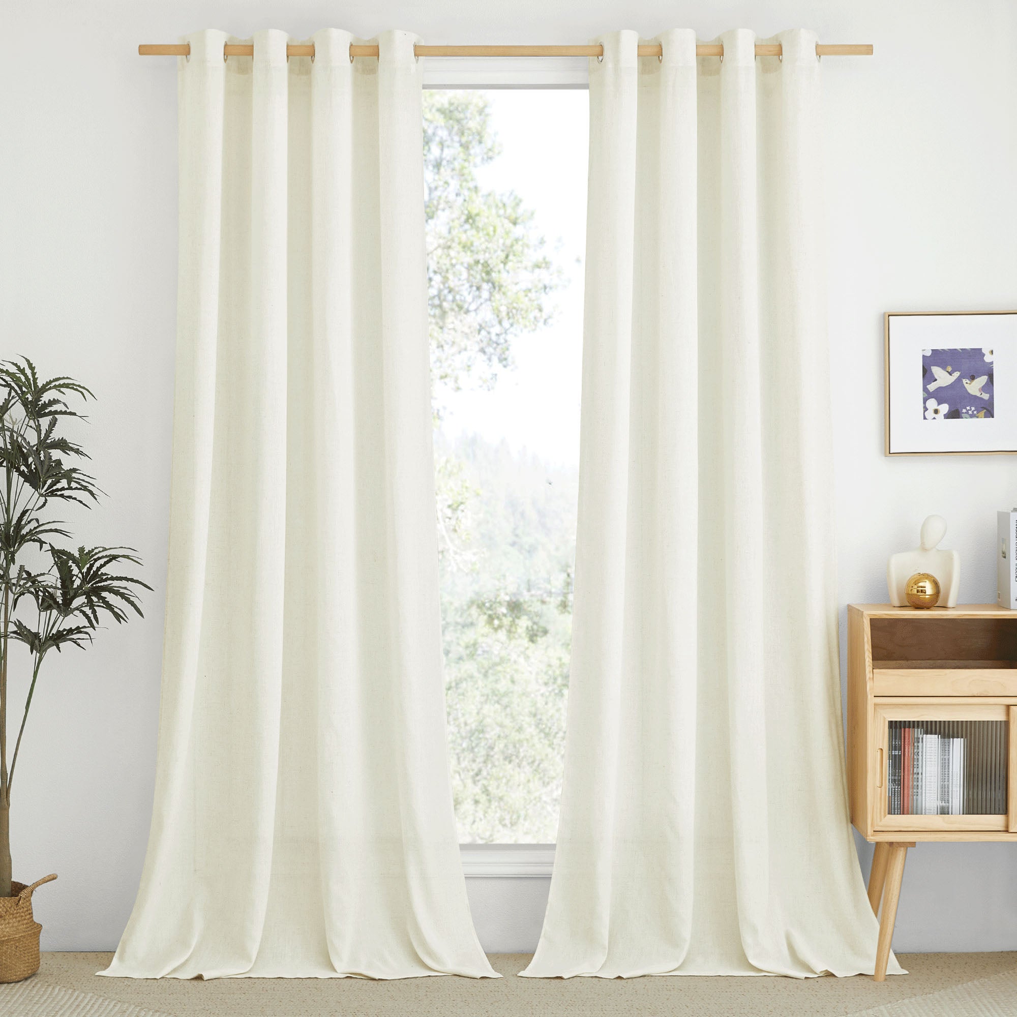 Full-length cream linen curtains with grommet tops hanging on a wooden rod in a bright, naturally lit room.
