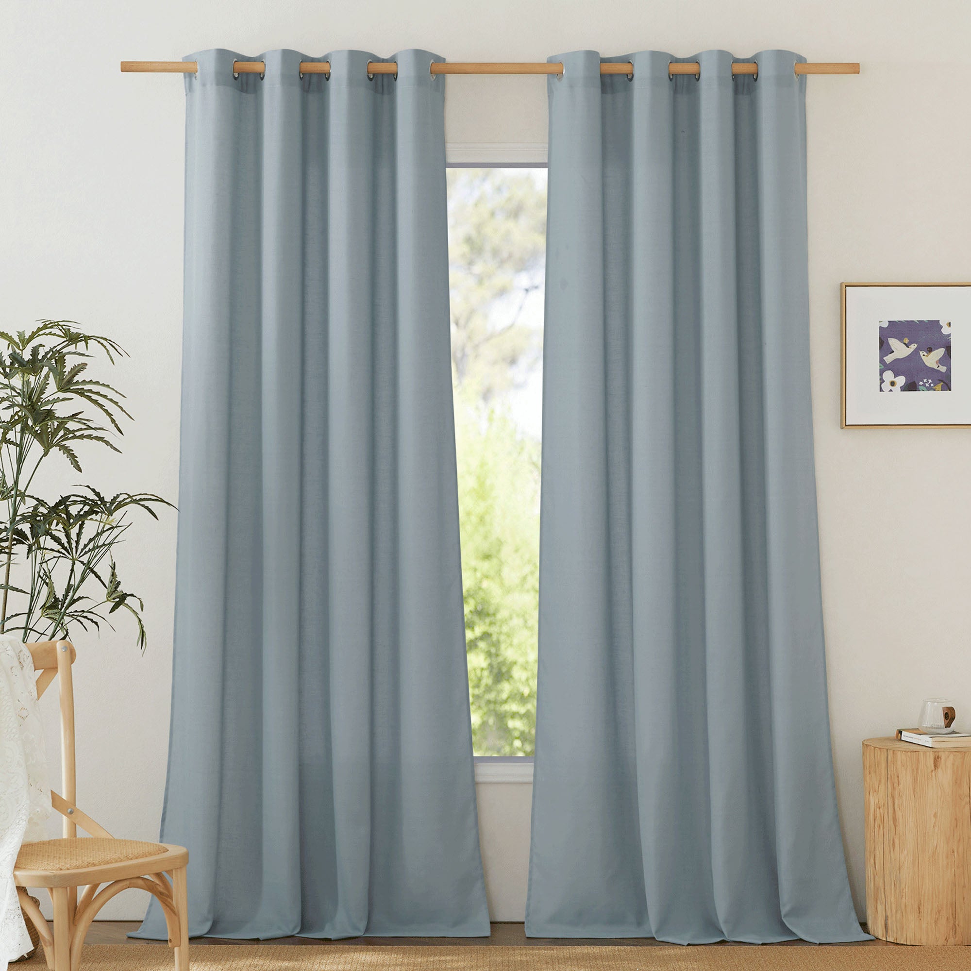 Two light blue linen curtain panels with silver grommets hang on a wooden rod, slightly parted over a window.