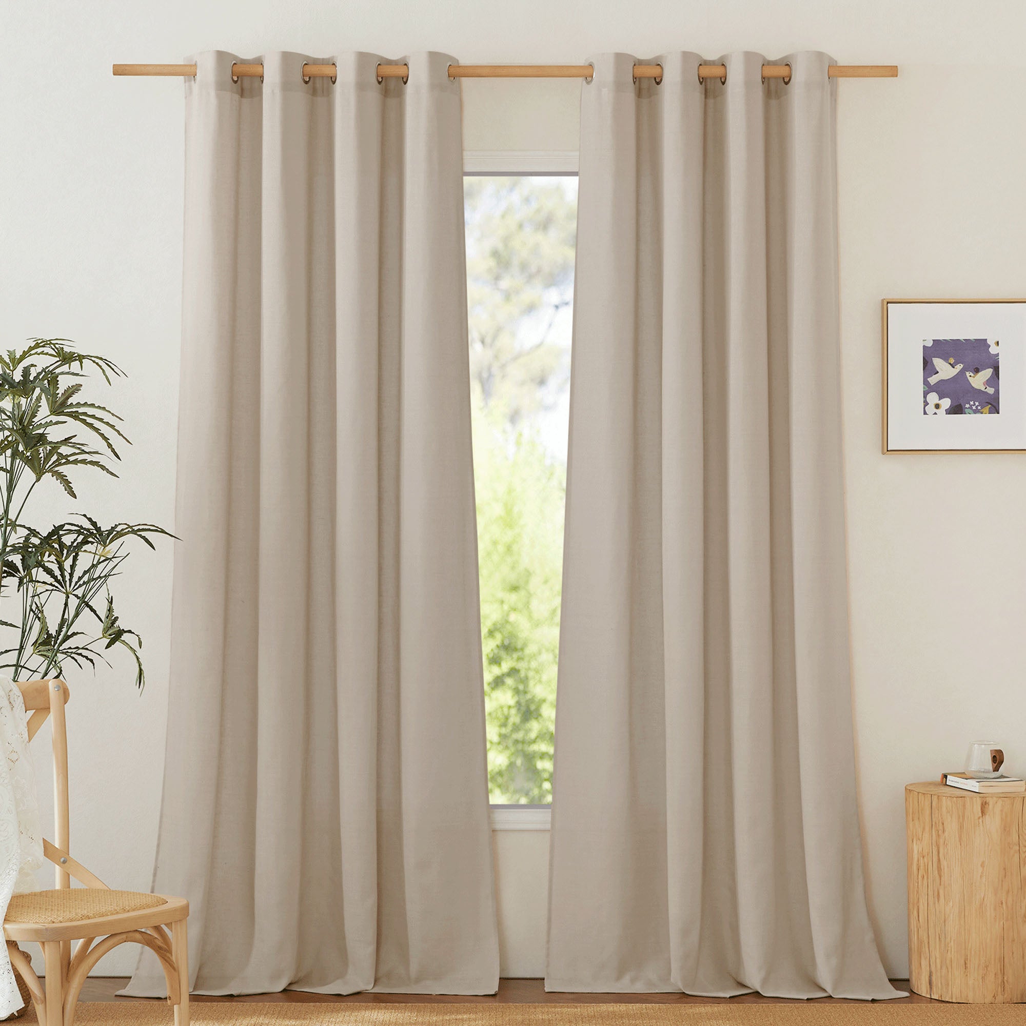 A pair of long, beige linen curtains with metal grommets hanging on a wooden rod over a window in a styled room.