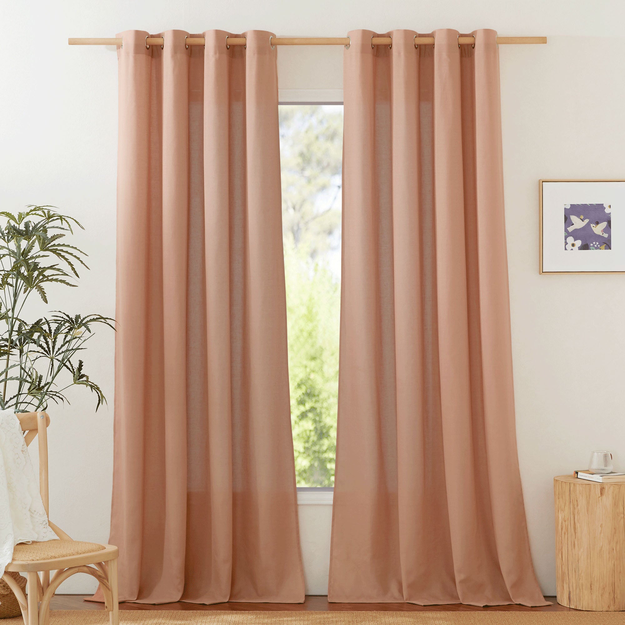 Dusty rose linen curtains with silver grommets hang on a wooden rod, framing a bright window in a styled room.