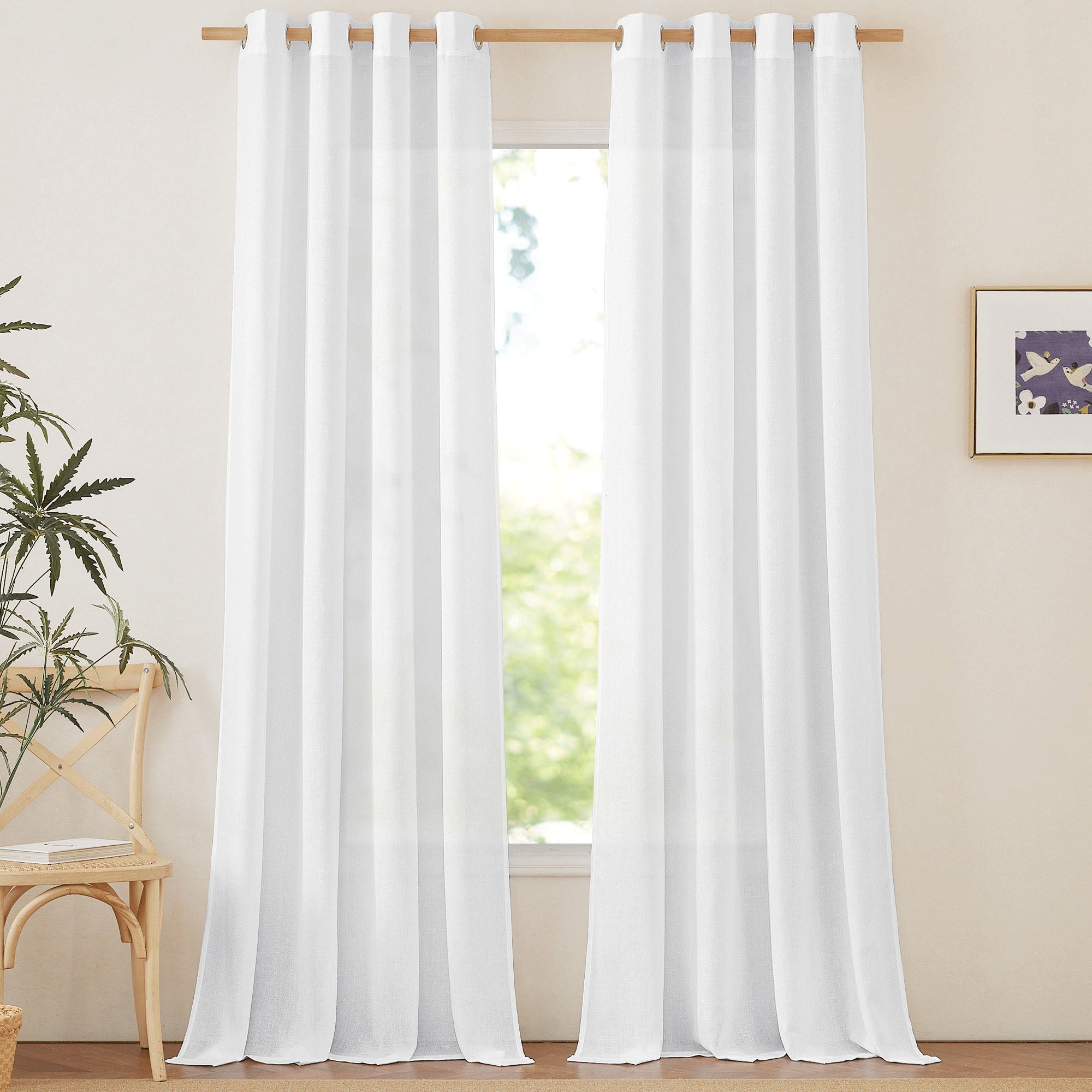 White semi-sheer linen curtains with grommets hanging on a wooden rod, framing a sunlit window.