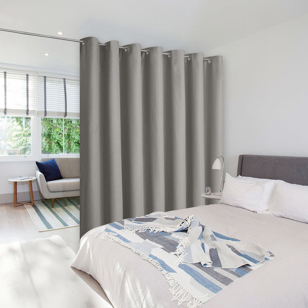 A grey room divider curtain on a tension rod separates a bedroom from a sitting area in a bright, modern room.