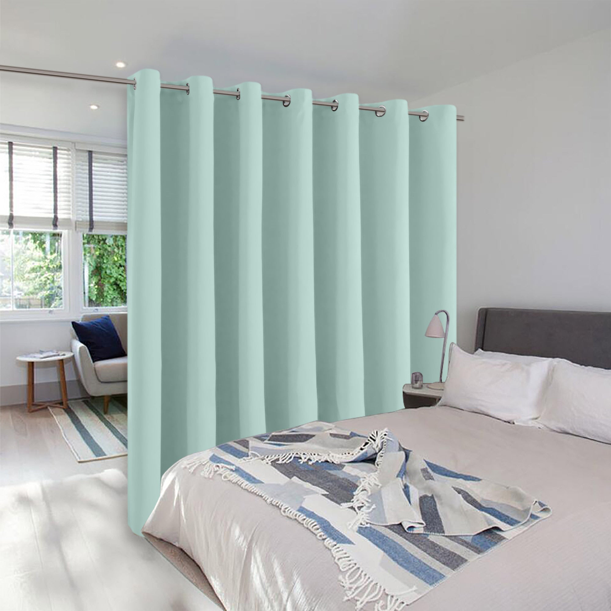 A light green room divider curtain with silver grommets hung on a ceiling rod to separate a bedroom from a small seating area.