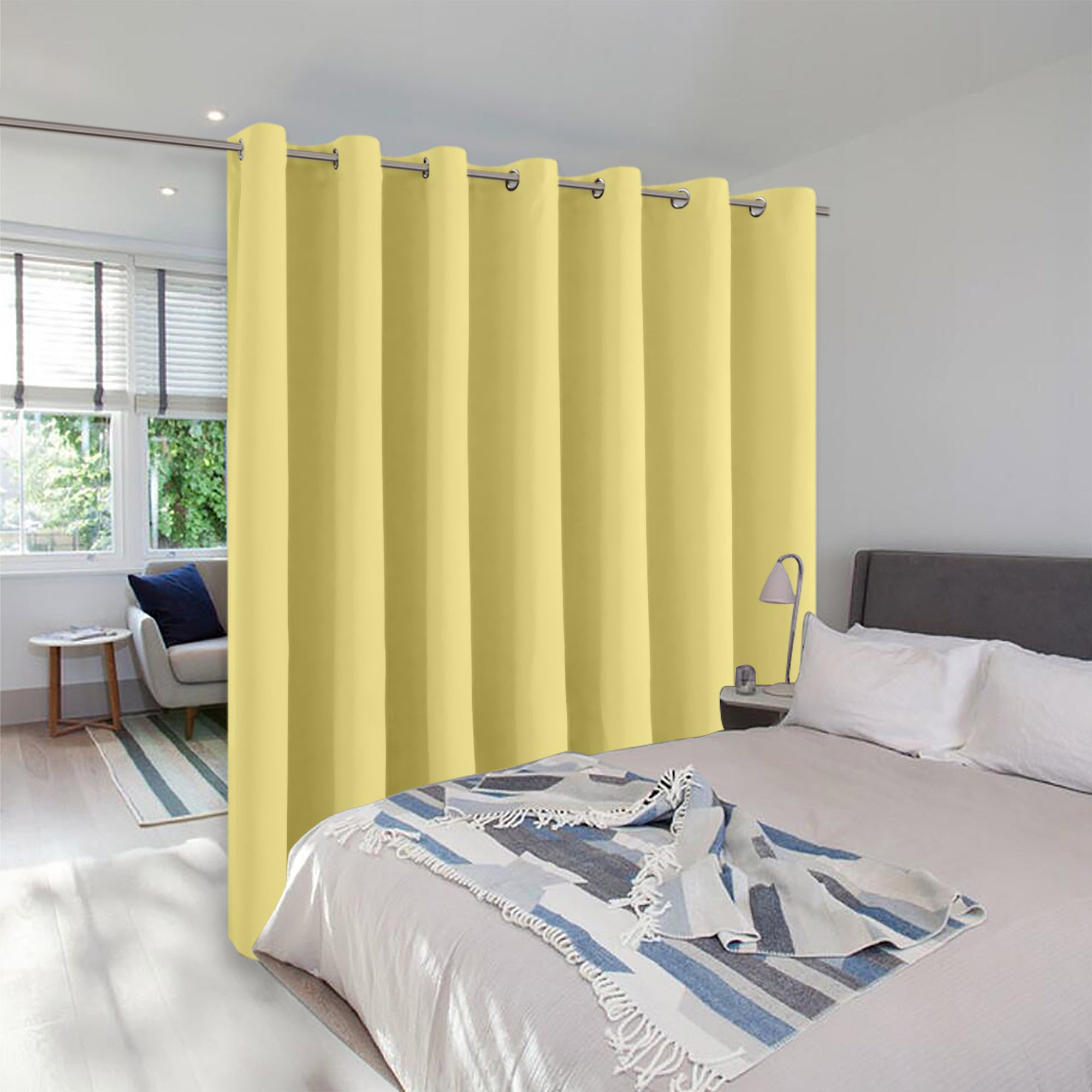 A pale yellow room divider curtain with silver grommets hangs to create privacy between a bed and a small sitting area.