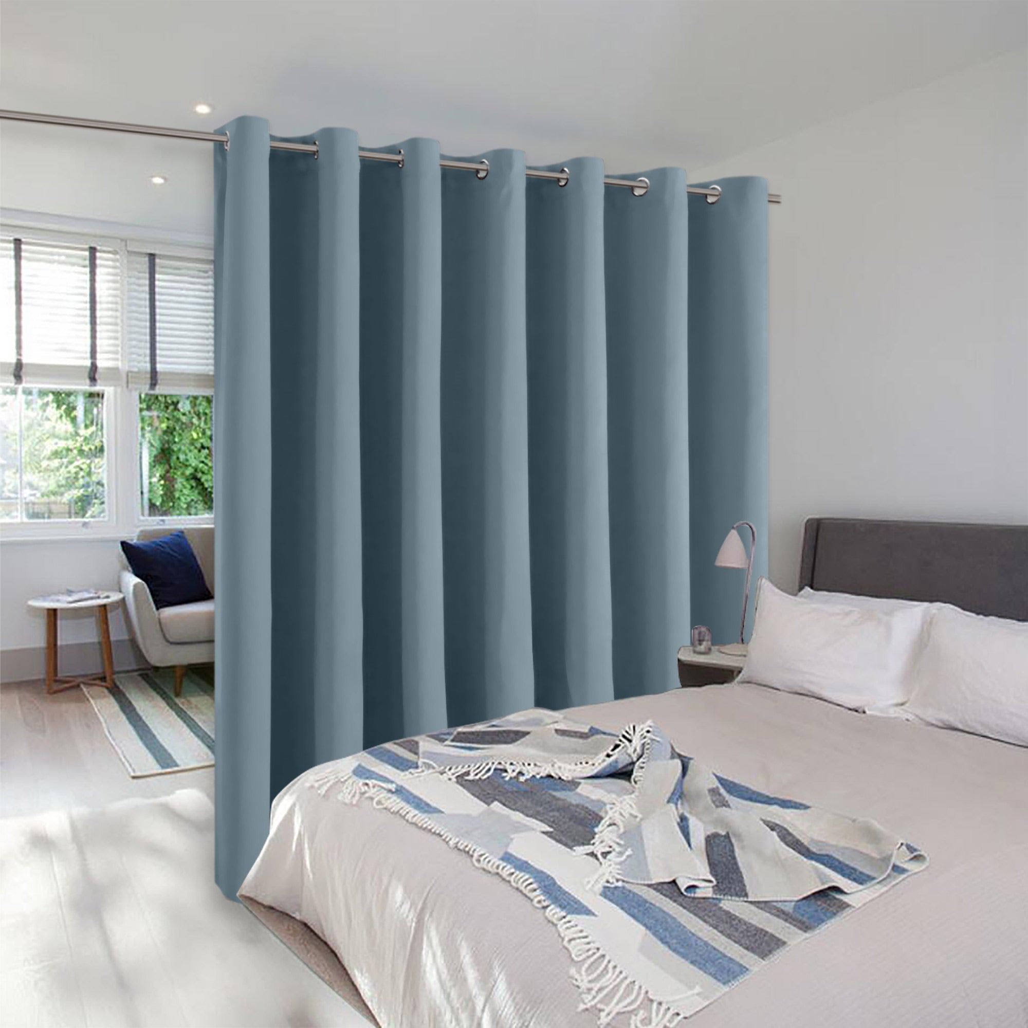 A dusty blue room divider curtain with grommets separates a bed from a sunlit sitting area in a modern bedroom.
