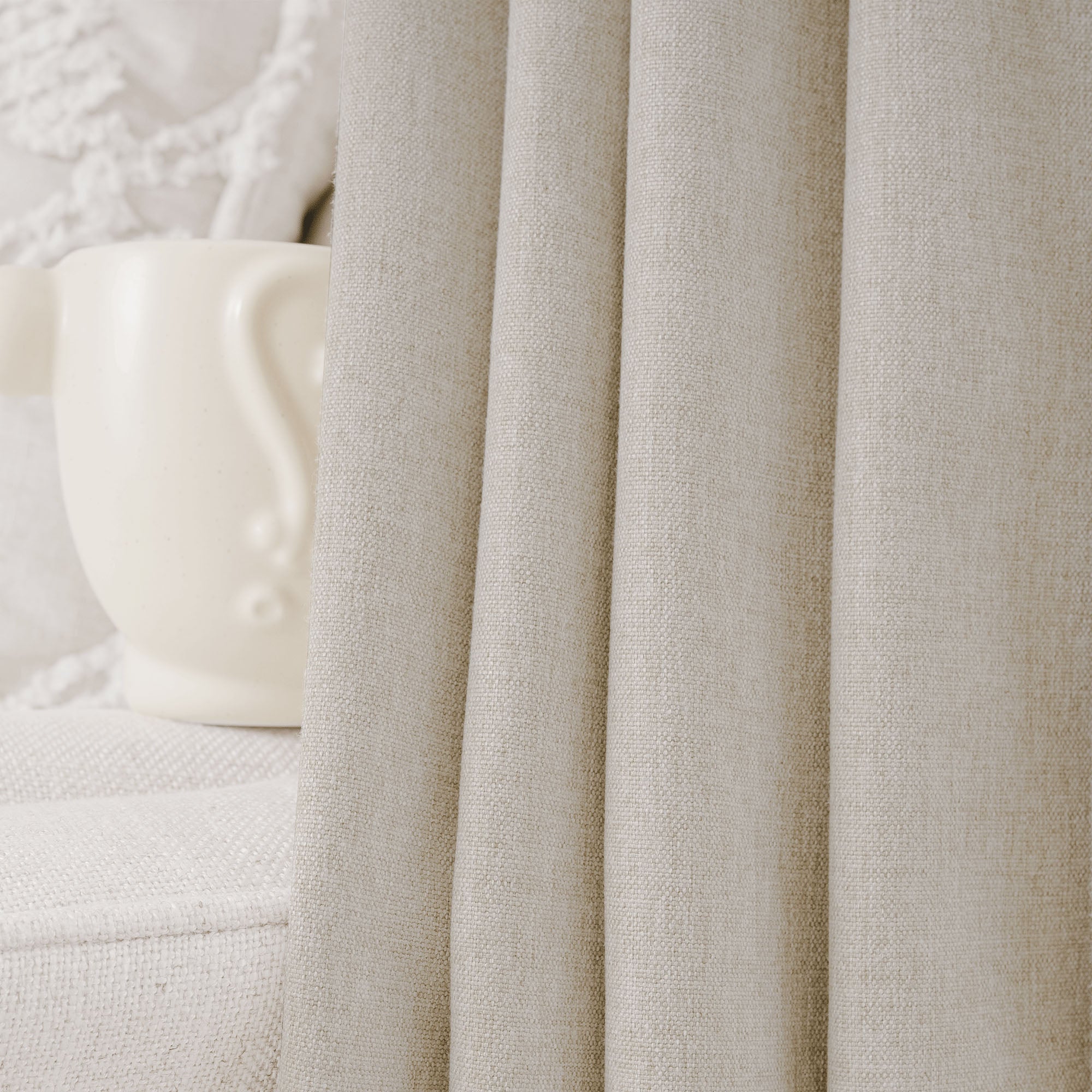 Detail view of soft beige cotton linen curtain, showcasing its natural texture and graceful folds. A hint of a white decorative object is visible.
