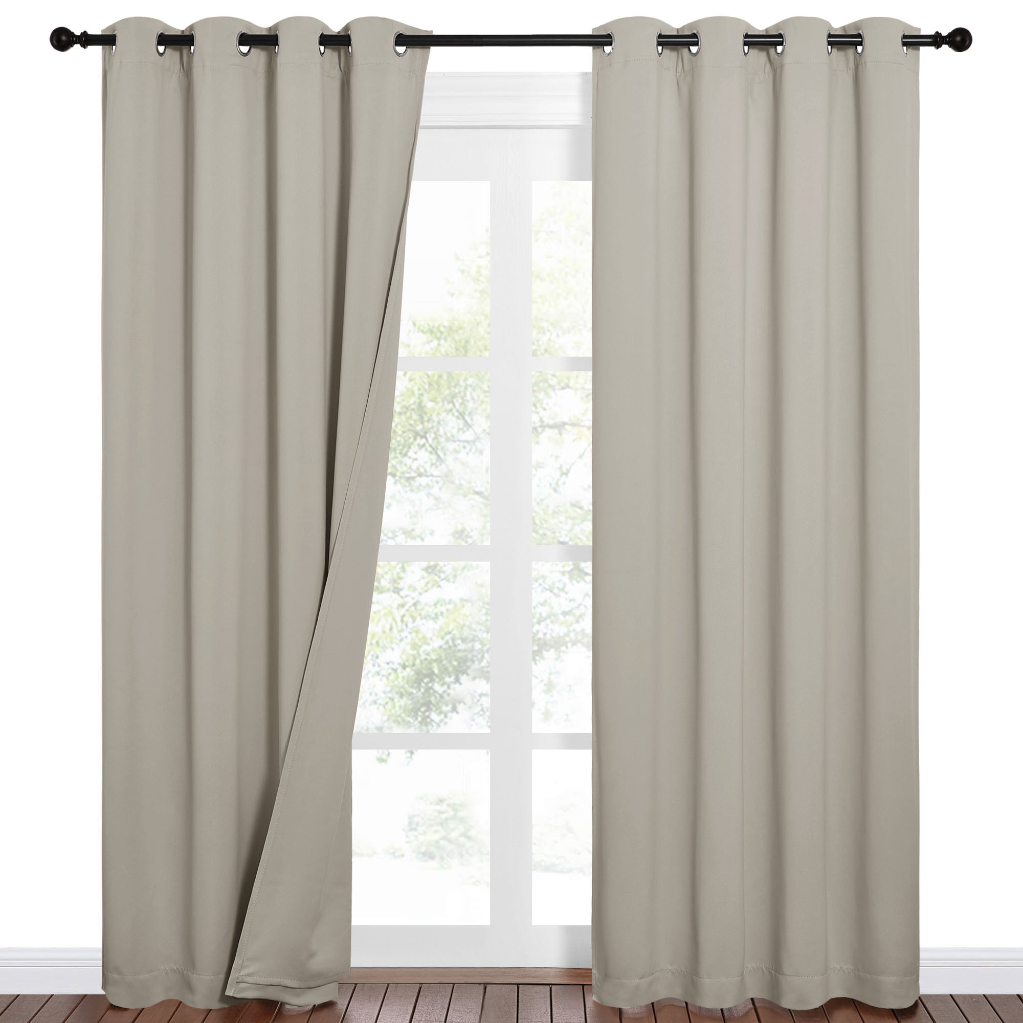Beige sound barrier 100% blackout curtains with grommet tops on a black rod, one partially open to show a window.