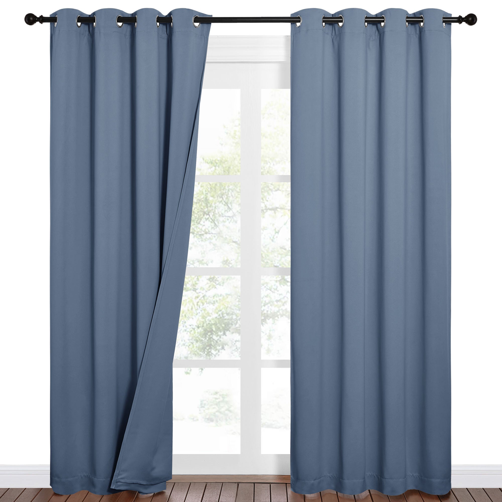Pair of blue Sound Barrier 100% blackout grommet curtains on a rod, slightly open, revealing a sunlit window.