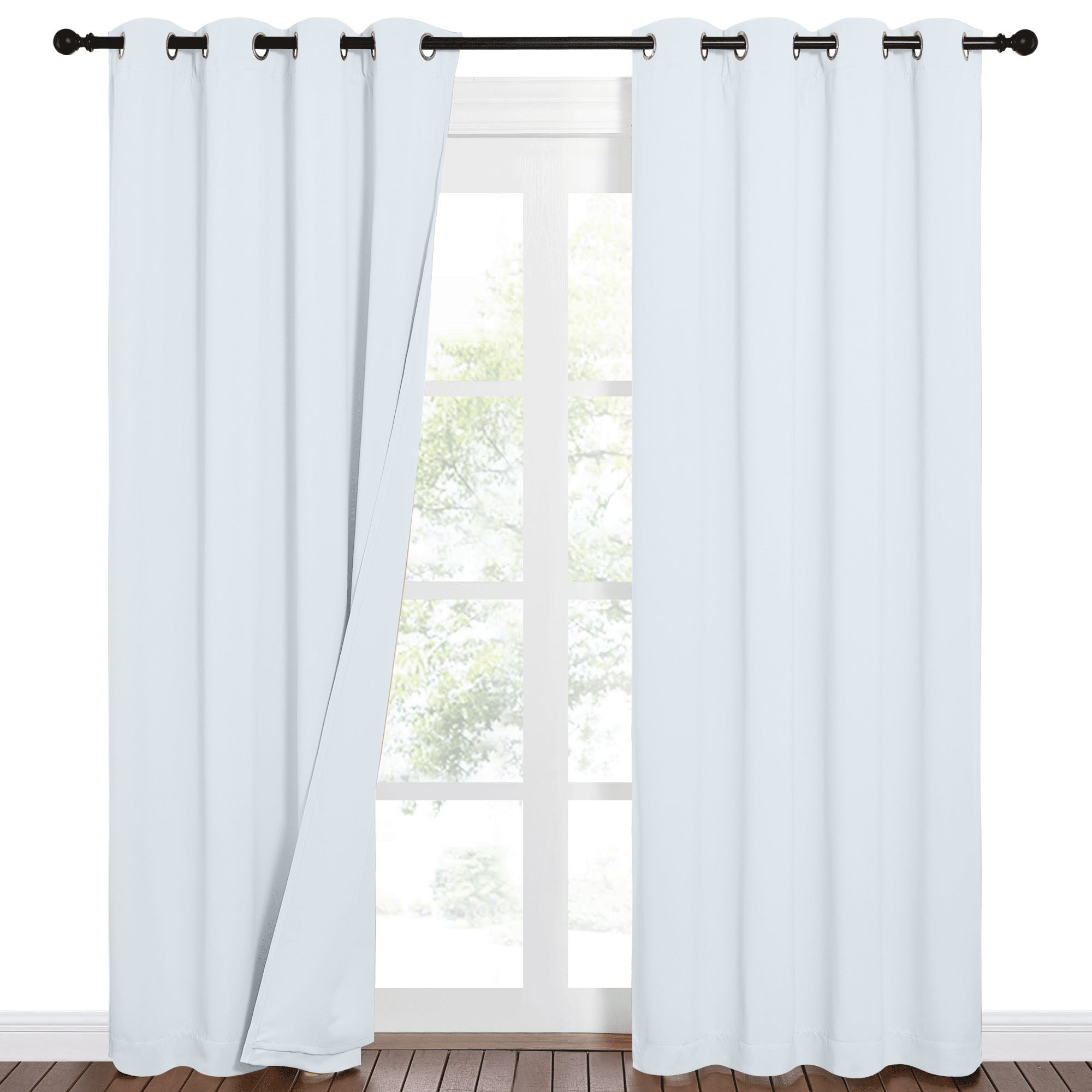 Light blue 100% blackout grommet curtains partially drawn, revealing a sunlit window with trees and a wood floor.