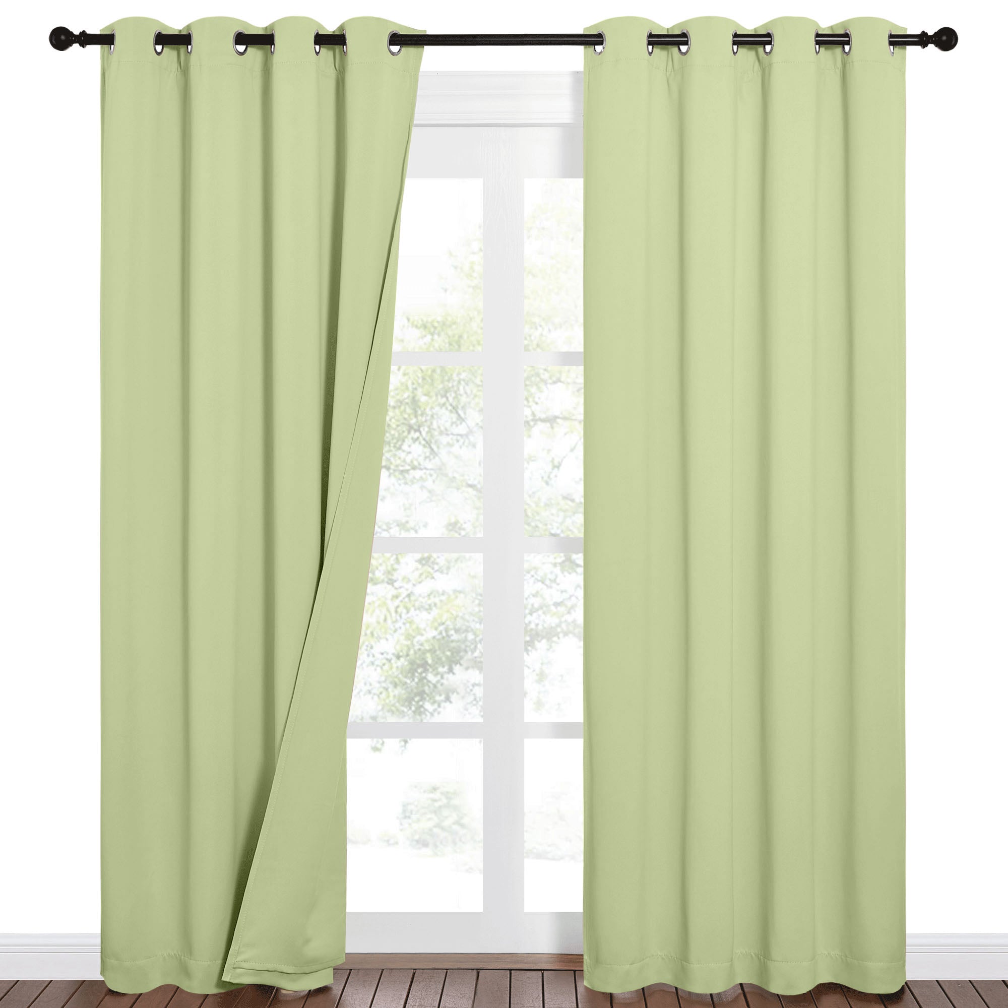 Pale green Sound Barrier 100% blackout grommet curtains on a dark rod. One panel is slightly open, revealing a bright window and outdoor trees.