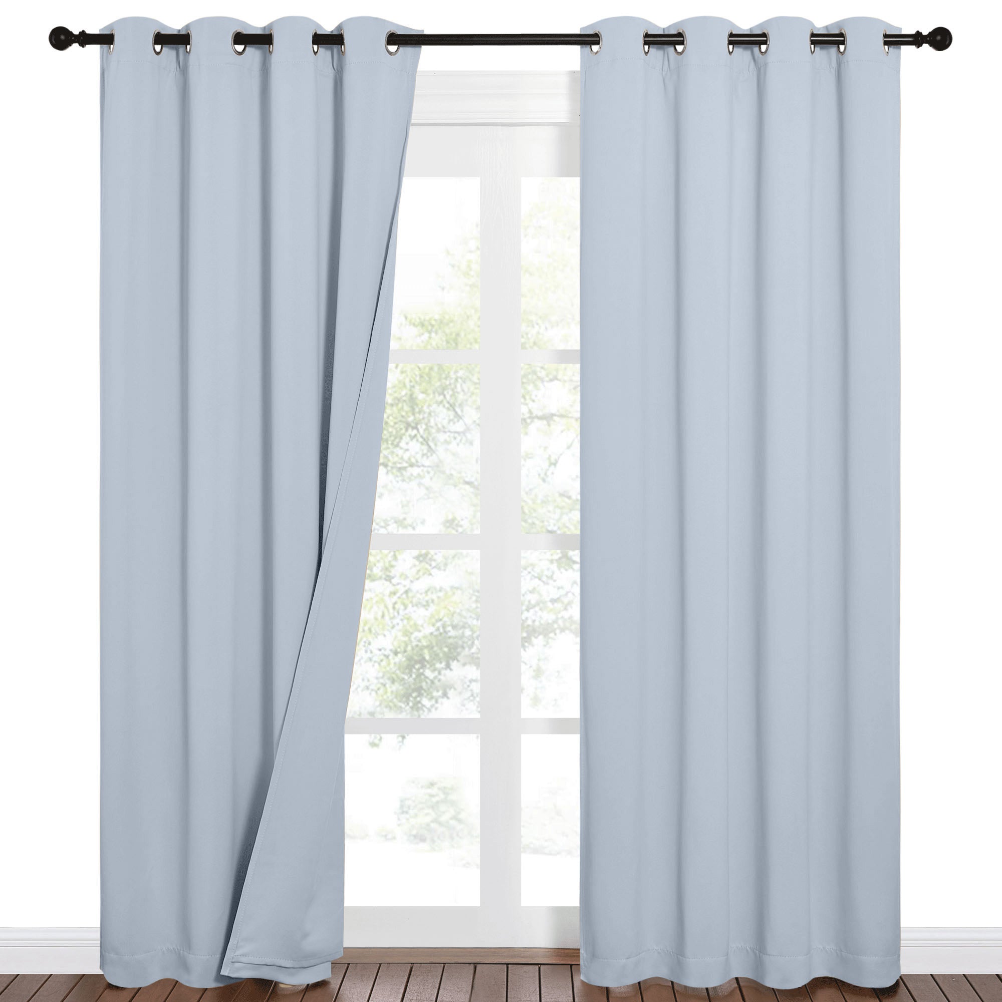 Light blue Sound Barrier 100% blackout grommet curtains on a dark rod, one panel open over a window with trees.