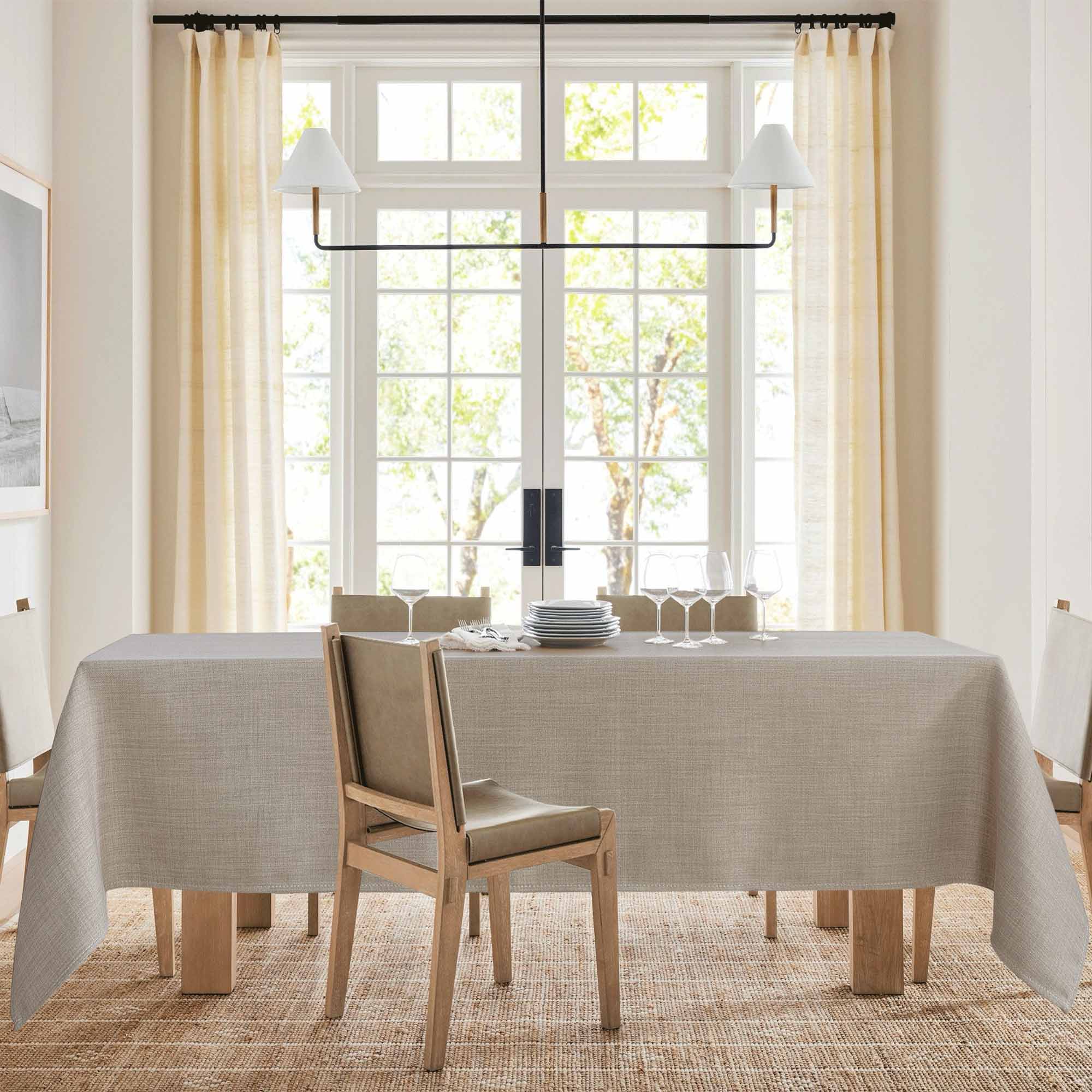 Tan textured tablecloth covering a dining table set with plates and wine glasses in a bright dining room with large windows.