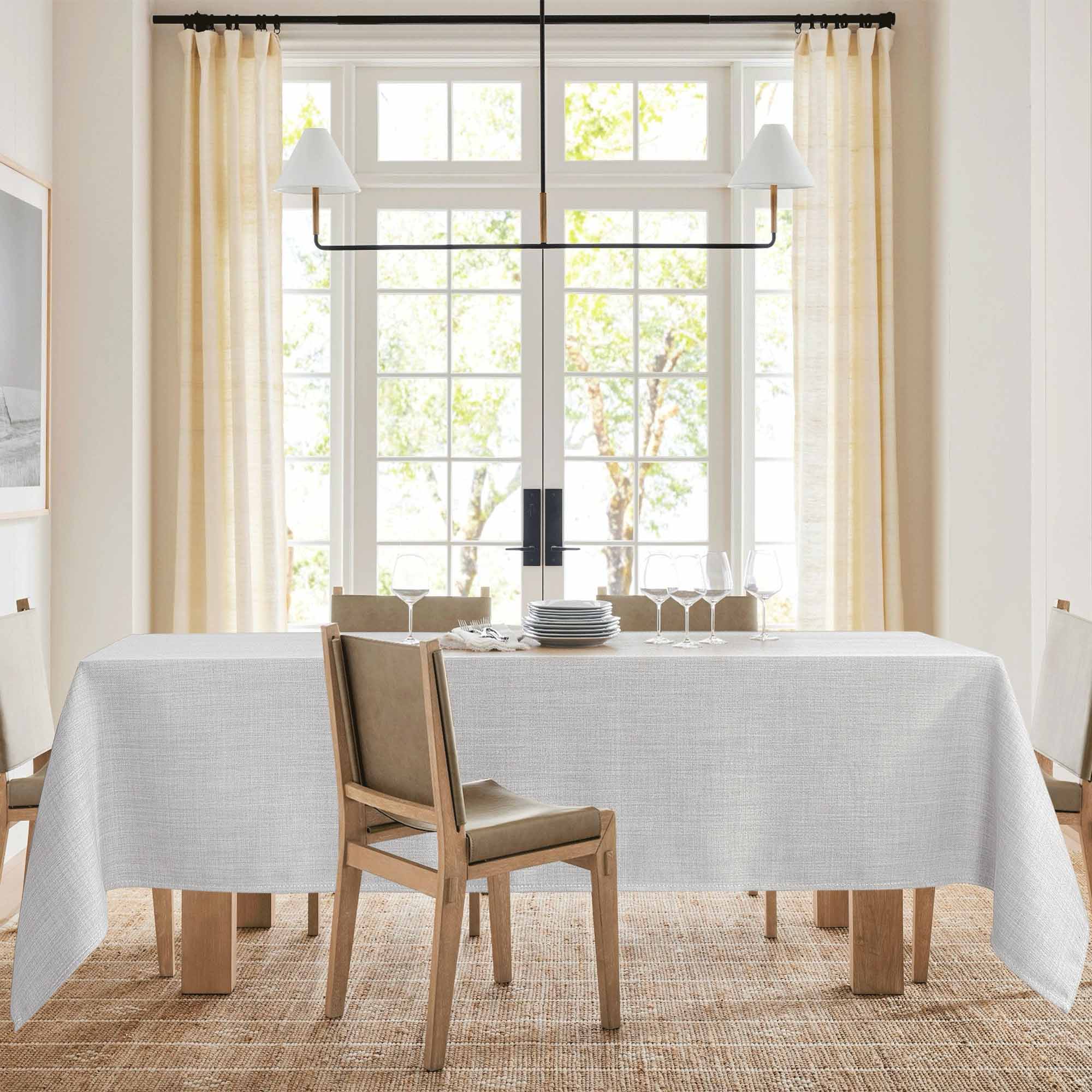 Textured light grey tablecloth covering a wooden dining table, set with plates and wine glasses in a bright dining room.