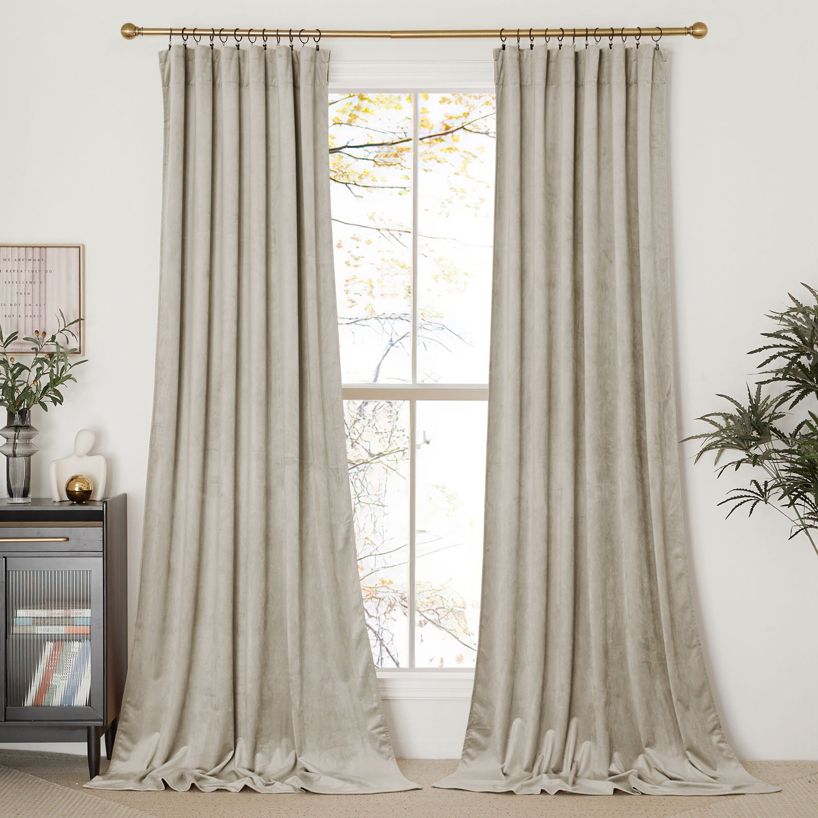 A pair of floor-length light beige velvet curtains hanging on a gold curtain rod, framing a tall window in a bright room.