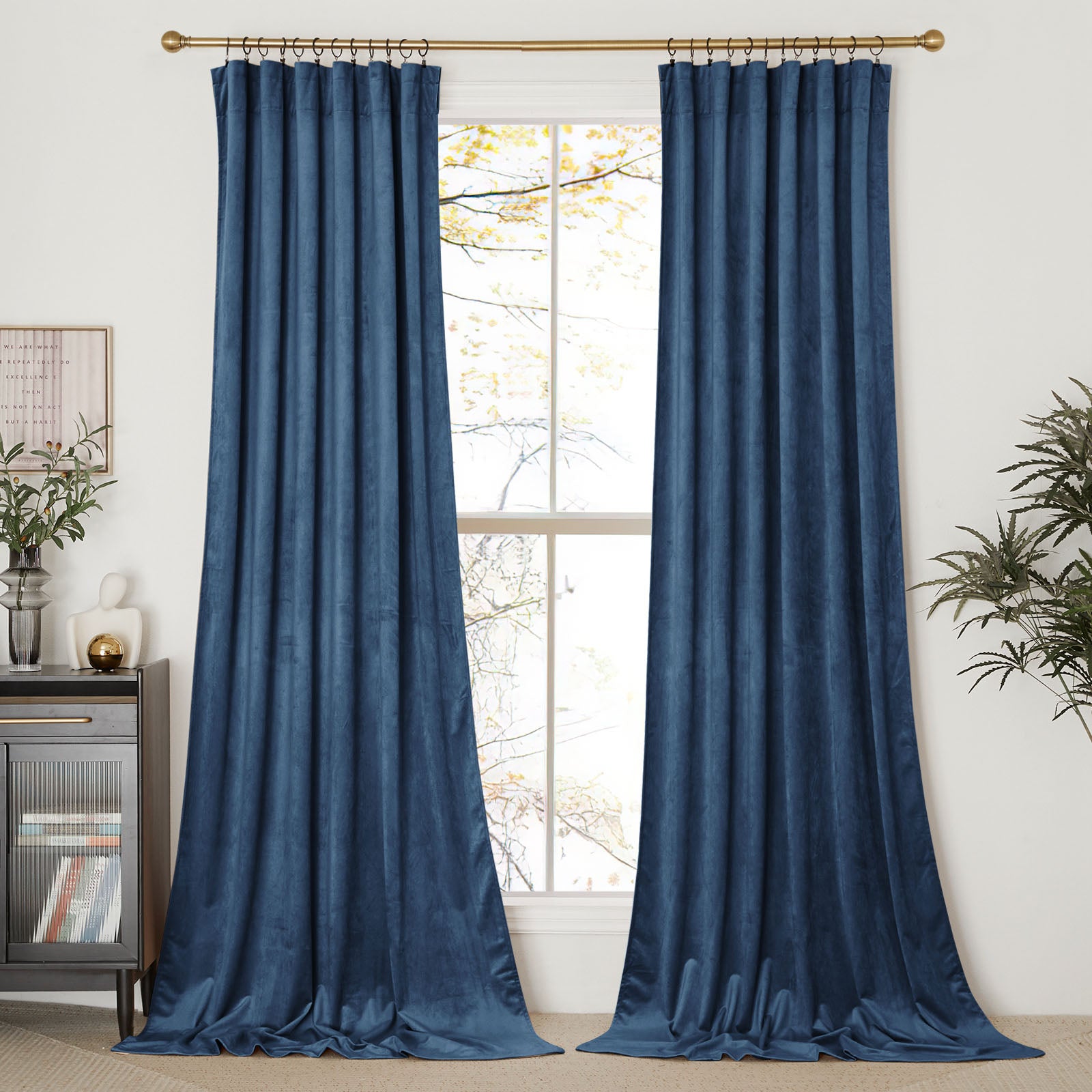 A pair of long, blue velvet curtain panels hanging on a gold curtain rod, framing a bright window in a styled room.