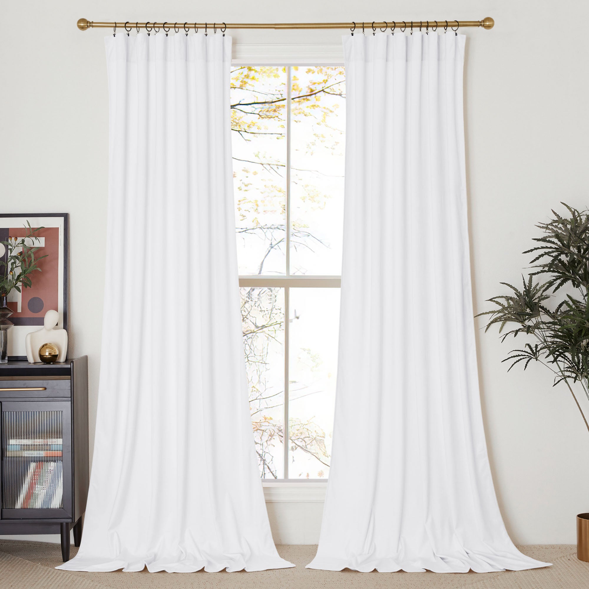Two long white velvet curtain panels hang from a gold rod, framing a sunny window in a decorated living room setting.