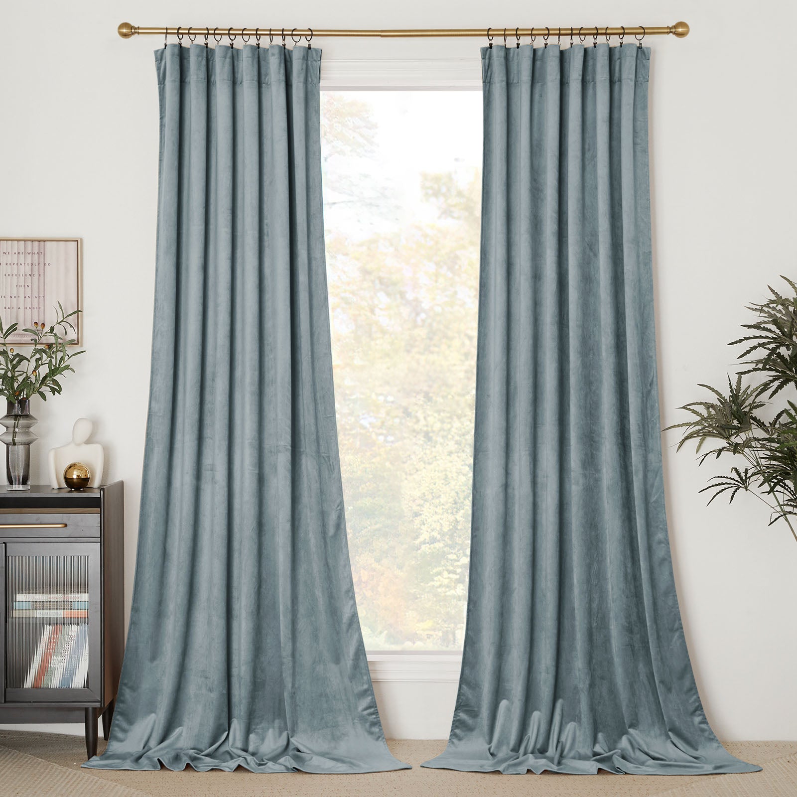 A pair of long, dusty blue velvet curtains hanging on a brass rod, framing a bright window in a modern living room.