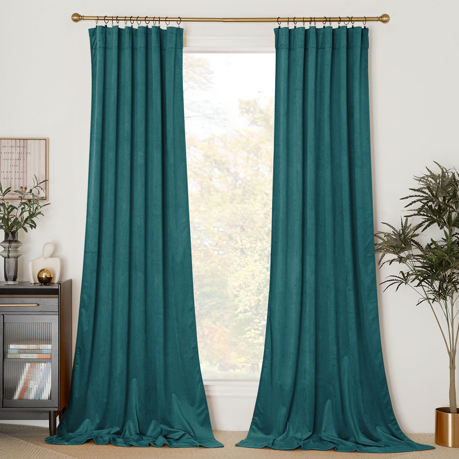 Two long teal velvet curtains with back tabs hanging on a gold rod with rings, framing a large window in a living room.
