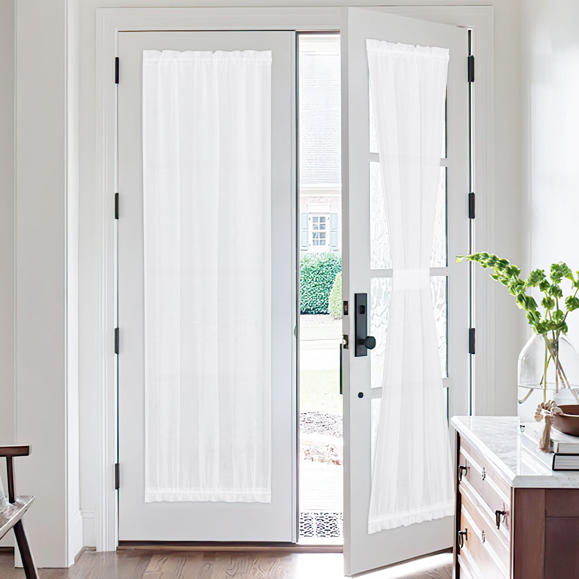 White sheer voile door curtains installed on bright French doors. One curtain tied back, revealing an outdoor garden.