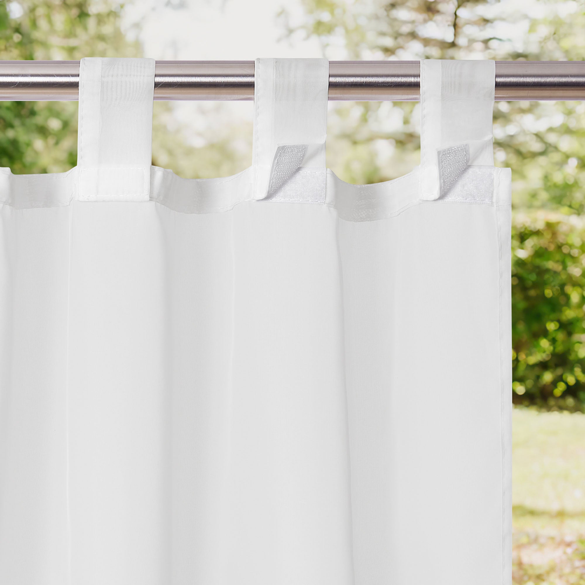 White waterproof sheer outdoor curtain with tab top Velcro closures on a silver rod, against a blurred green garden.