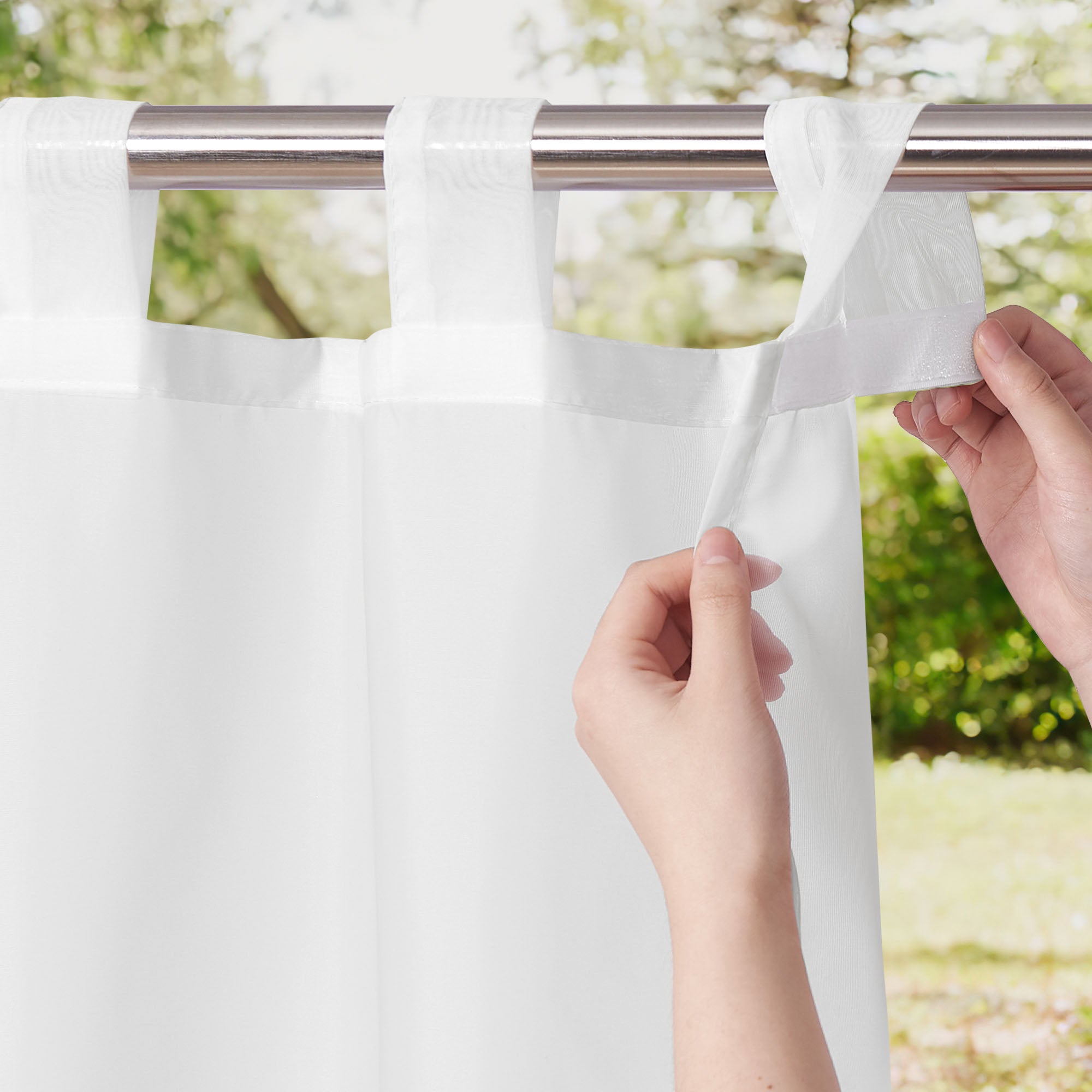 Hands fastening the tab top of a white waterproof sheer outdoor curtain to a silver rod, showing the hook-and-loop closure detail.