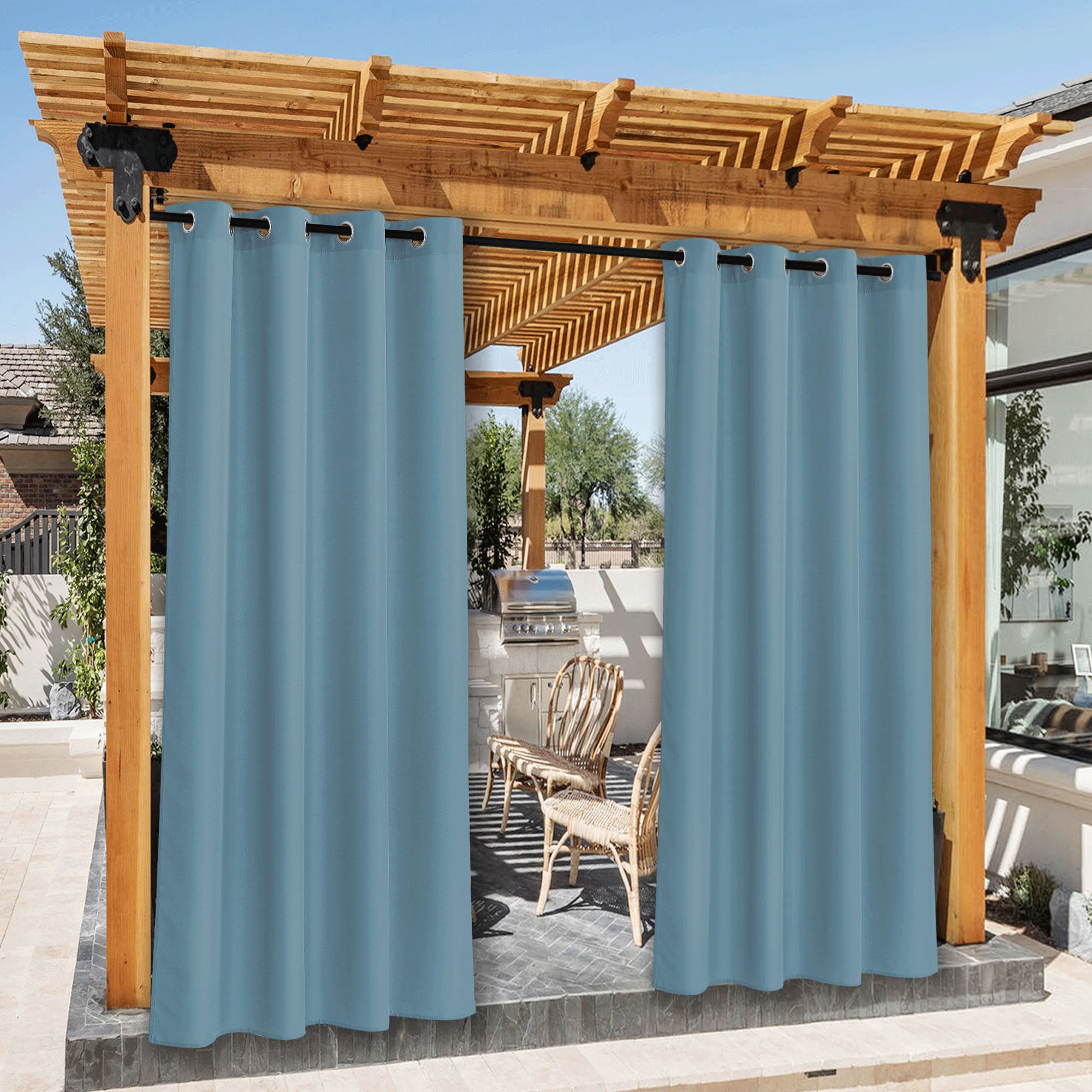Blue waterproof canvas outdoor curtains on a wooden pergola, revealing a patio with a built-in grill and wicker chairs.