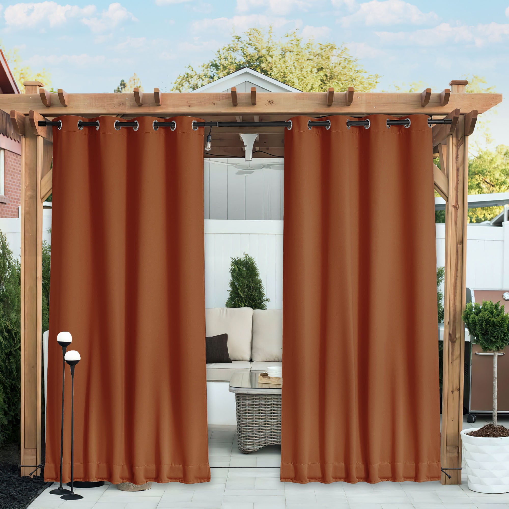 Rust-colored waterproof blackout outdoor curtains open on a wooden pergola, revealing a patio lounge area with a sofa.