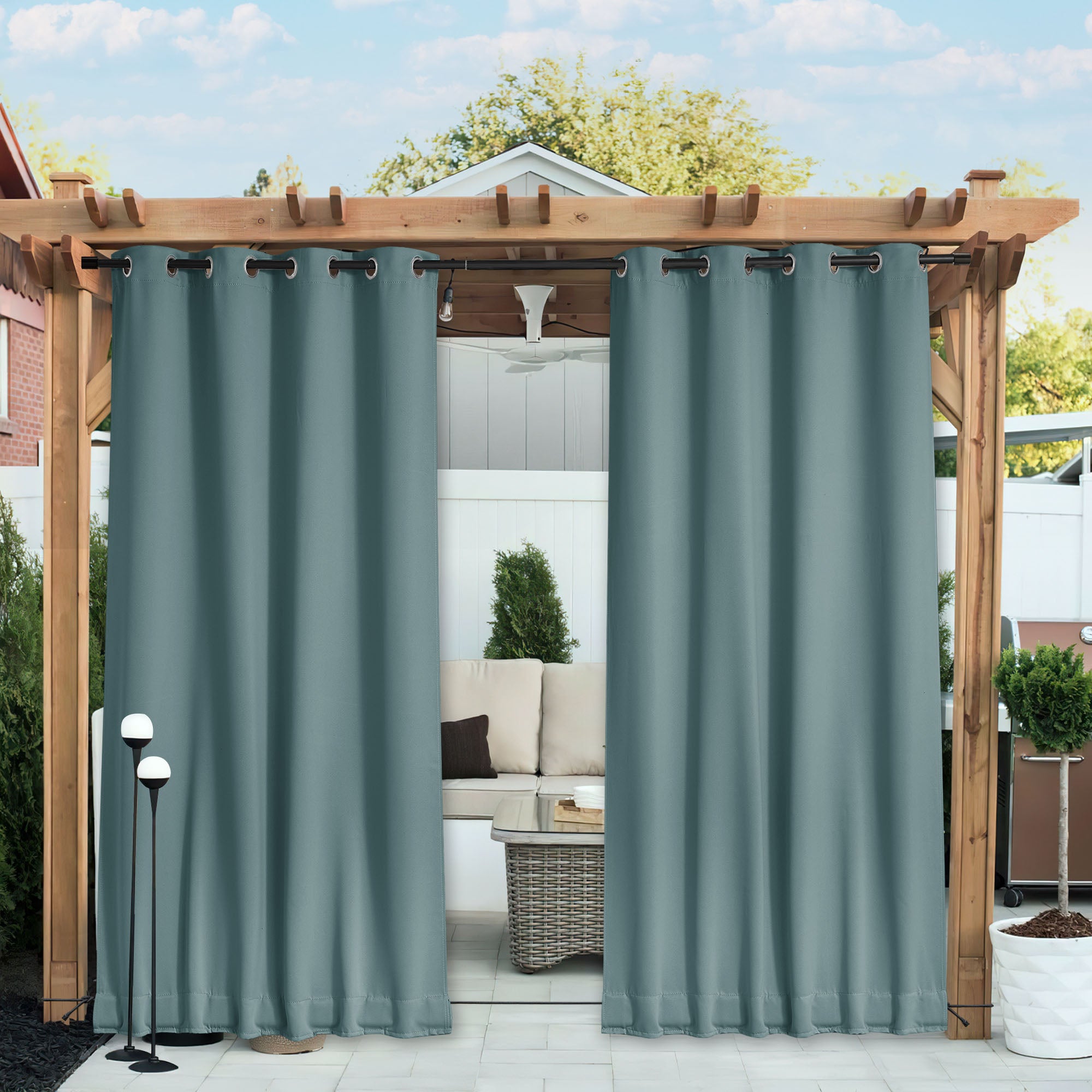 Two slate blue waterproof outdoor blackout curtains with grommet tops hanging from a pergola, revealing a patio lounge behind.