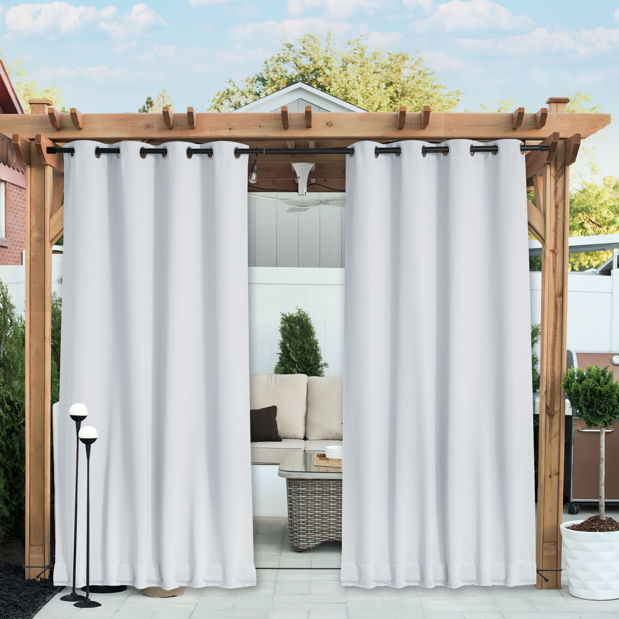Two white waterproof blackout outdoor curtains hanging from a wooden pergola, partially open to reveal a patio seating area.
