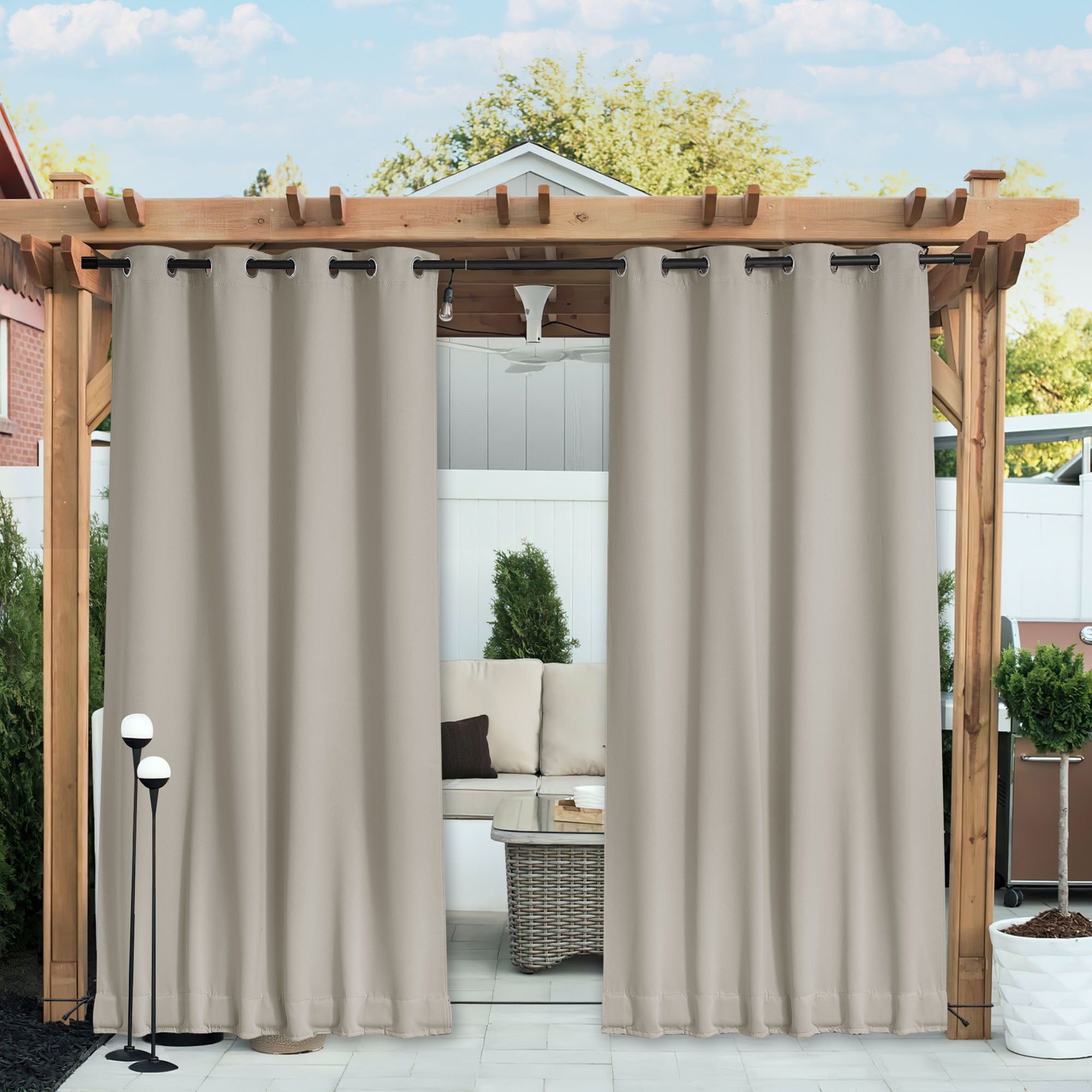 Two beige waterproof blackout outdoor curtains hanging from a wooden pergola, partially open, revealing a patio seating area.