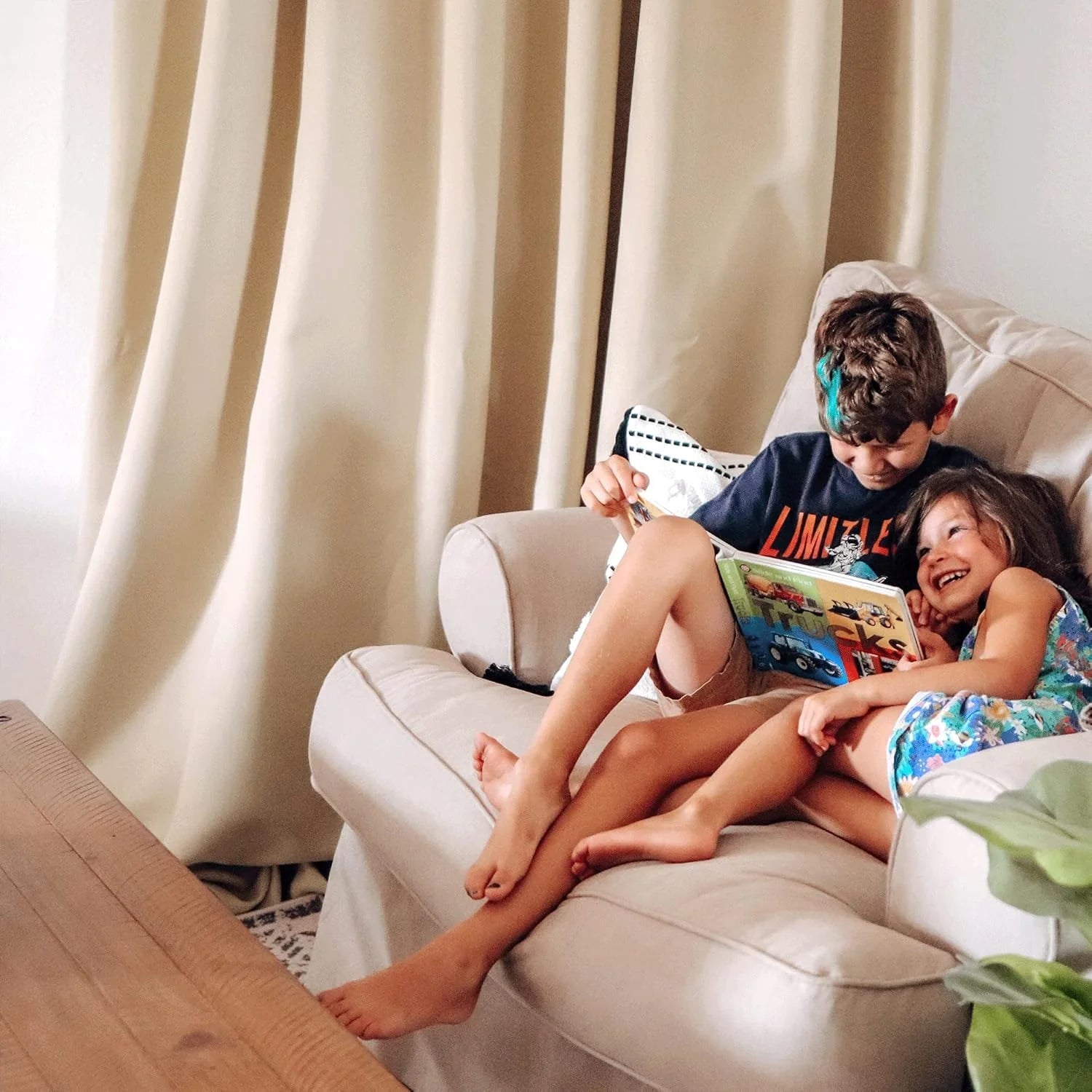 Two children reading together on a white armchair beside beige curtains