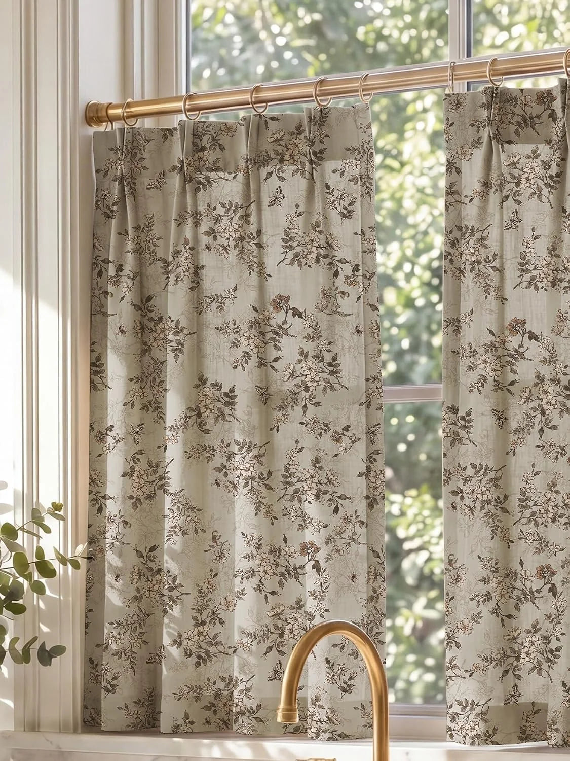 Beige botanical cafe curtains with brass rings hanging in a bright kitchen window