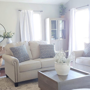 Beige fabric sofa with rolled arms, detailed nailhead trim, and dark wooden feet, styled in a bright living room.